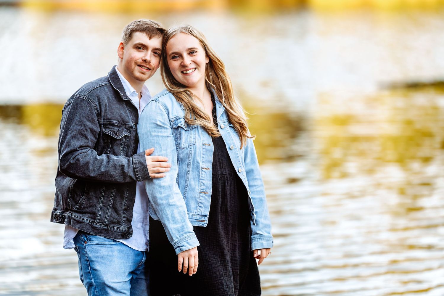 Couple poses together by waterfront with golden sunlight reflecting on lake surface.