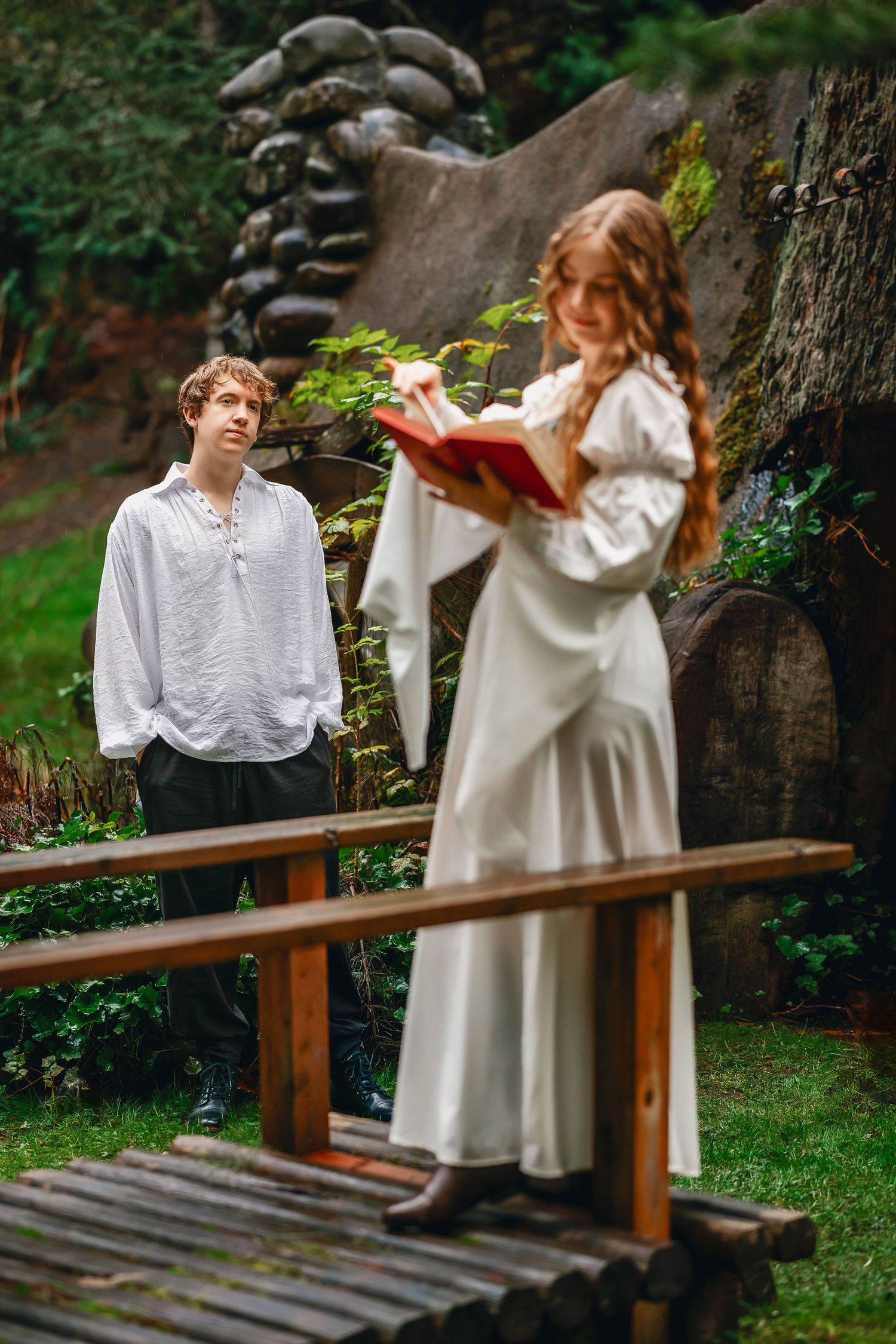 Two people in vintage white clothing stand on a wooden bridge in a lush garden setting.