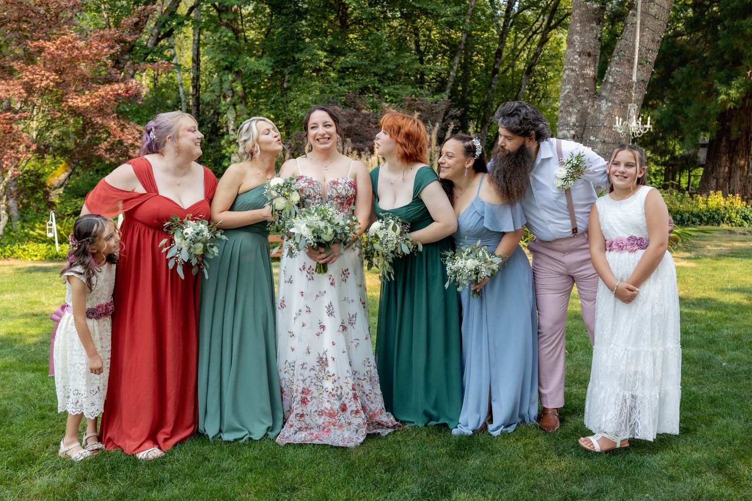 Wedding party in mix-and-match pastel and jewel-toned dresses poses together on green lawn during outdoor celebration.