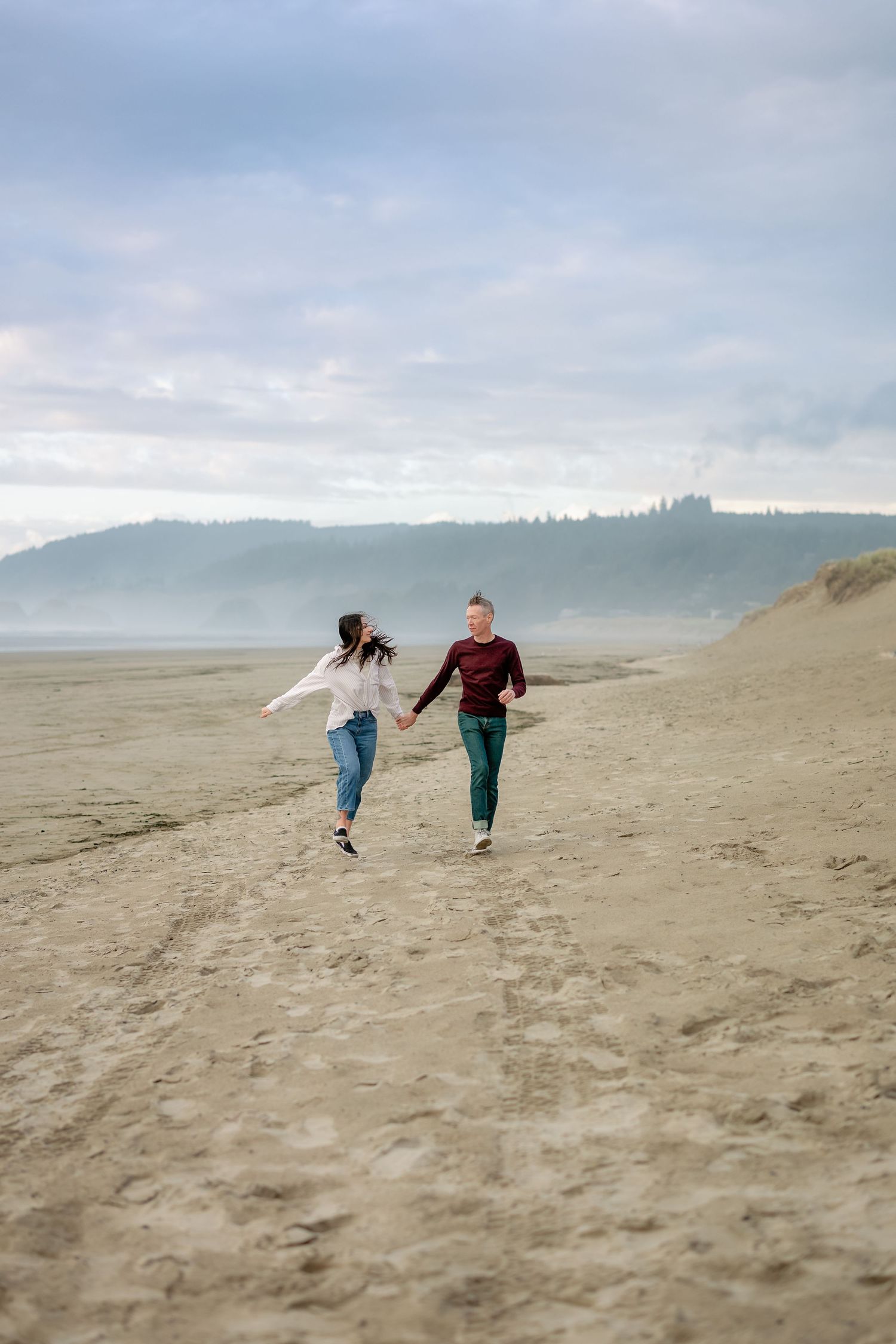 People run and play together on a misty beach with rolling sand dunes and hills in the background.