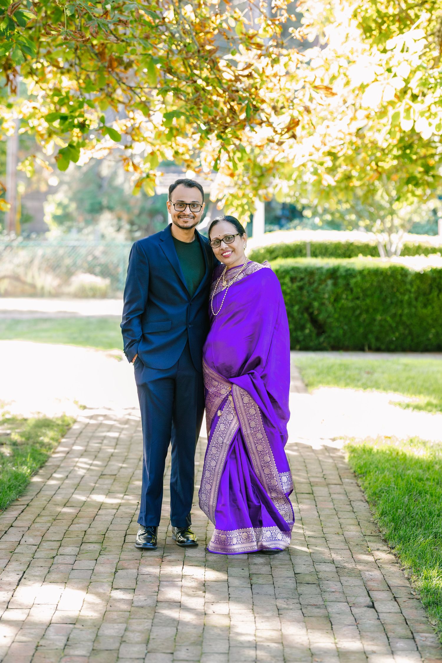 A couple stands together on a brick pathway in a garden setting, with one wearing a vibrant purple outfit.