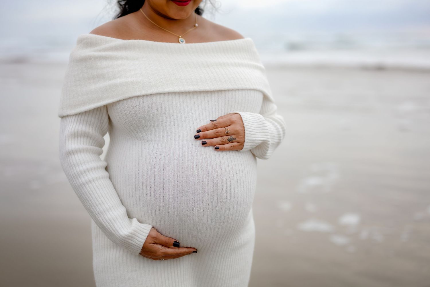 A pregnant woman in a white ribbed sweater poses on a beach during a maternity photoshoot.
