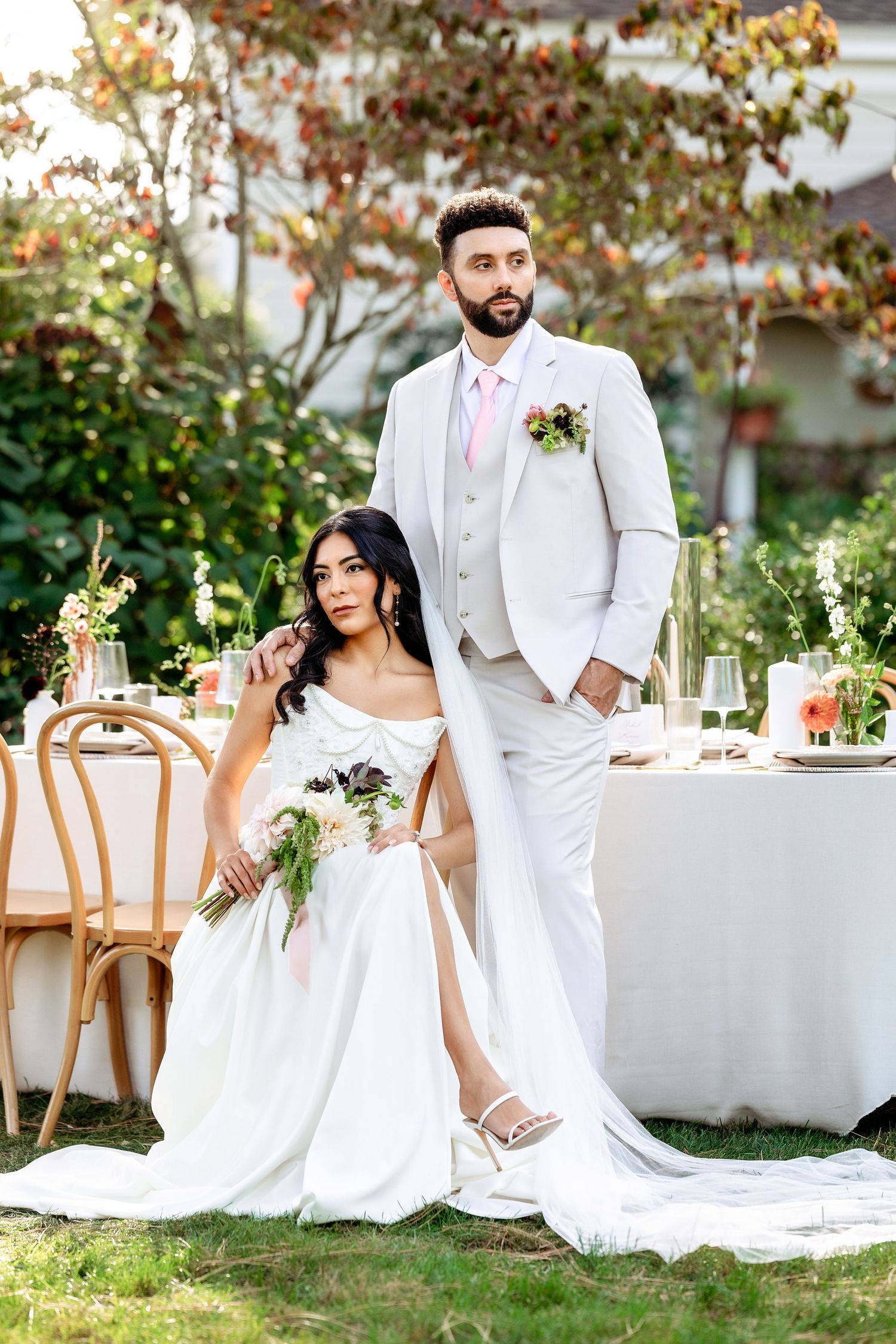 Elegant wedding couple poses on lawn with white table settings and floral decor in outdoor garden setting.