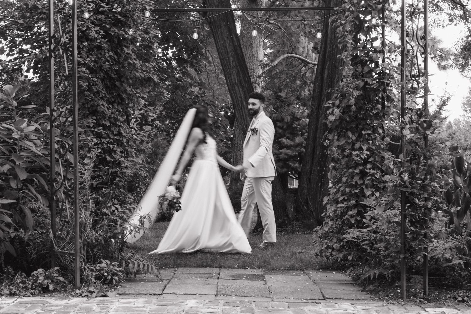 Black and white wedding photo series showing couple walking through tree-lined garden path.