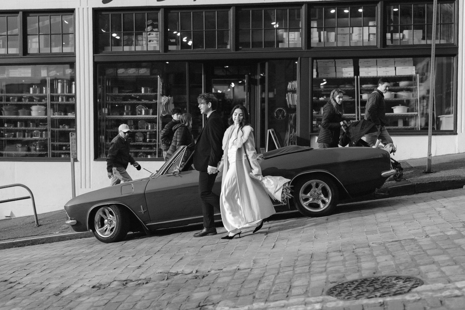 Black and white photo of couple walking on cobblestone street with parked cars and storefronts.