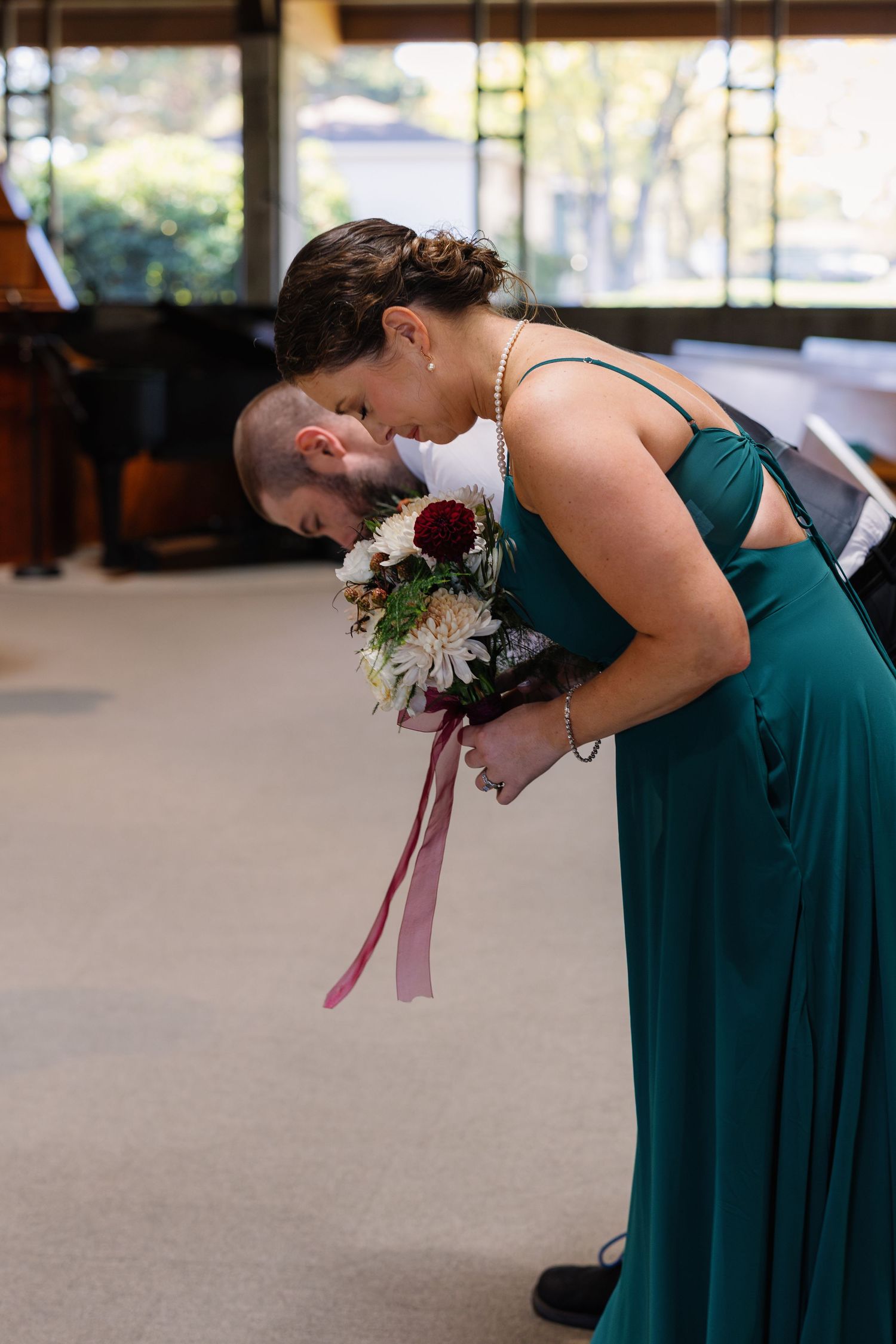 A bridesmaid in an emerald green dress bends down while holding a bouquet with burgundy ribbons at a wedding ceremony.