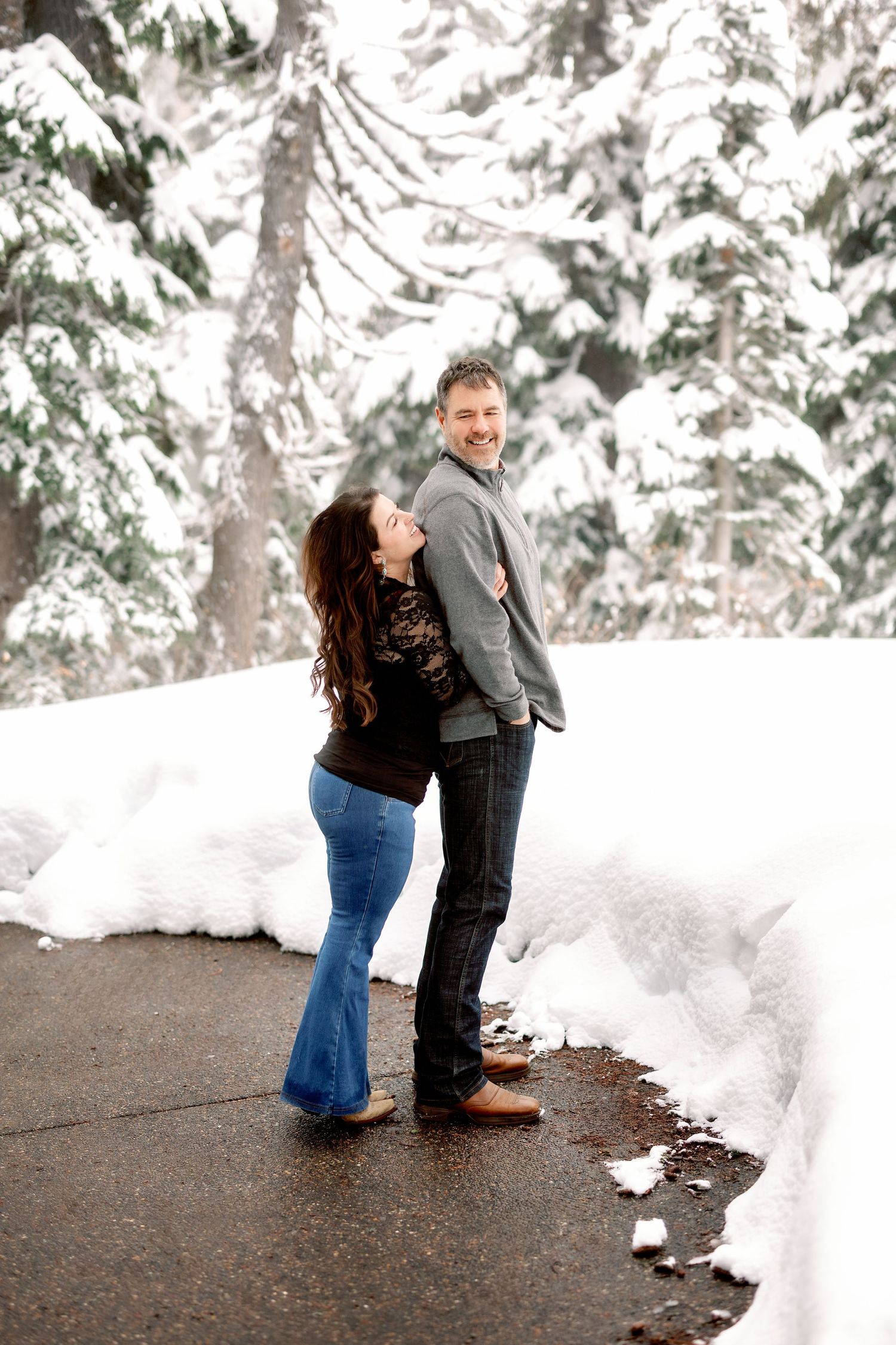 A couple shares a tender moment in a snowy winter forest setting with snow-covered evergreen trees in the background.