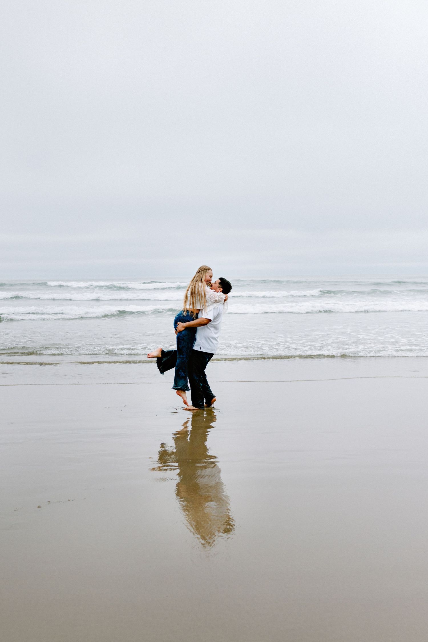 Couple embracing and kissing on a wet sandy beach with ocean waves in the background on an overcast day.