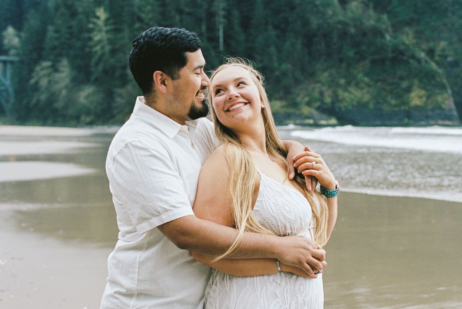 Happy couple embracing on a beach near a river, woman smiling up at man in white shirt with forest behind them.