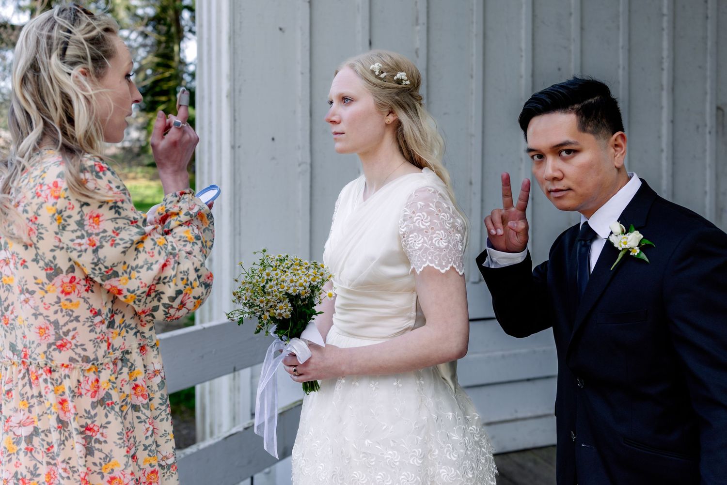 A wedding coordinator adjusts details while the bride holds a daisy bouquet and groom signals with his hand.