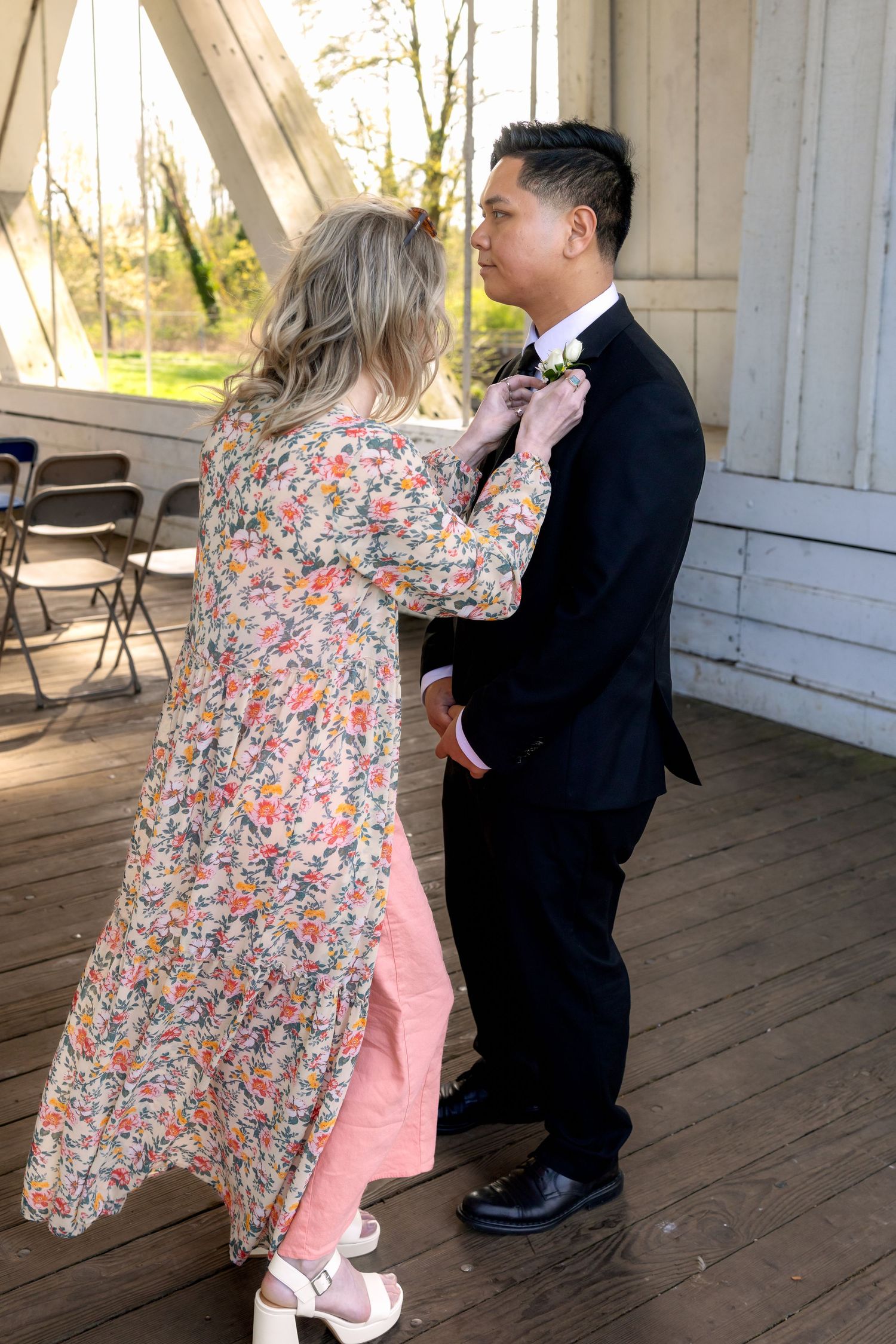 A woman in a floral dress adjusts the boutonniere on a groom in a black tuxedo at an outdoor wedding venue.