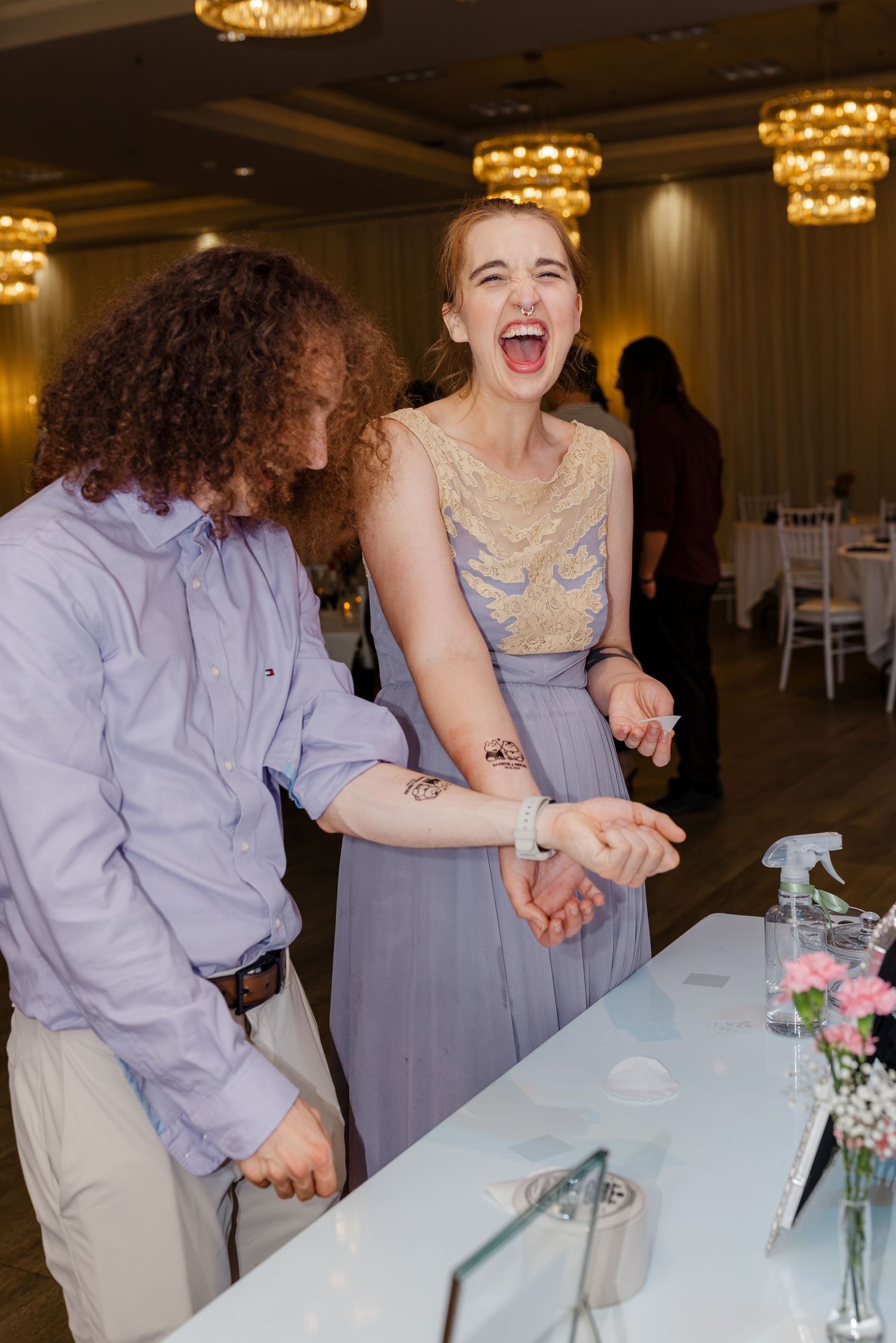 Woman in lavender gown laughing joyfully beside a curly-haired man at a wedding reception table.