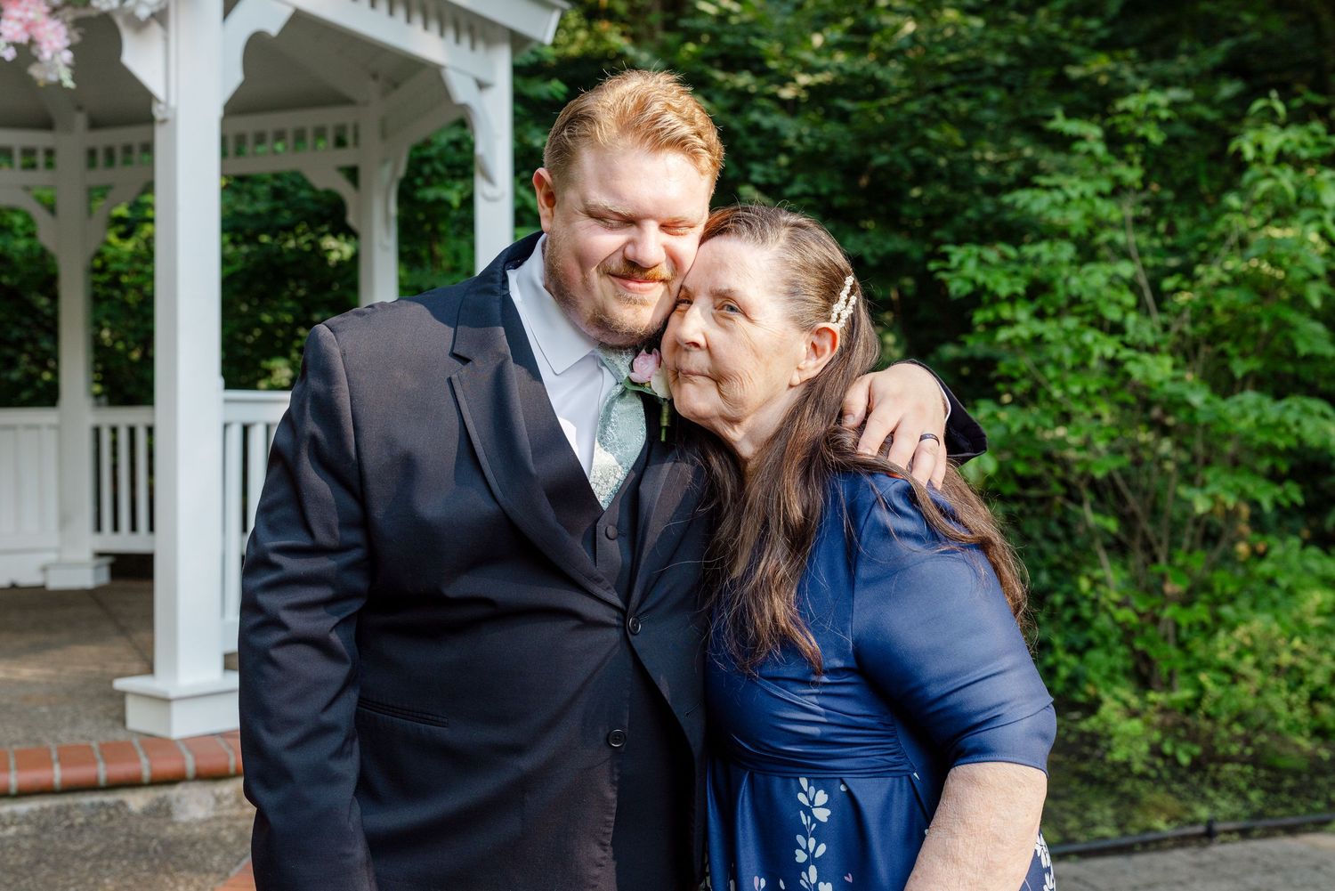 Redheaded groom in navy suit embraces older woman in blue dress near white gazebo outdoors.