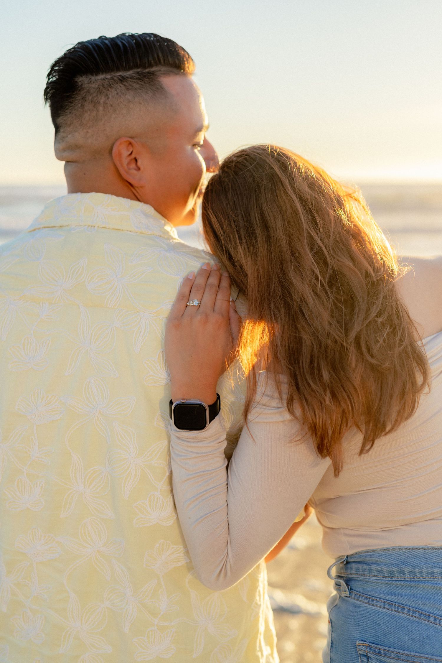Couple sharing intimate moment at beach sunset, woman with engagement ring embracing man from behind.