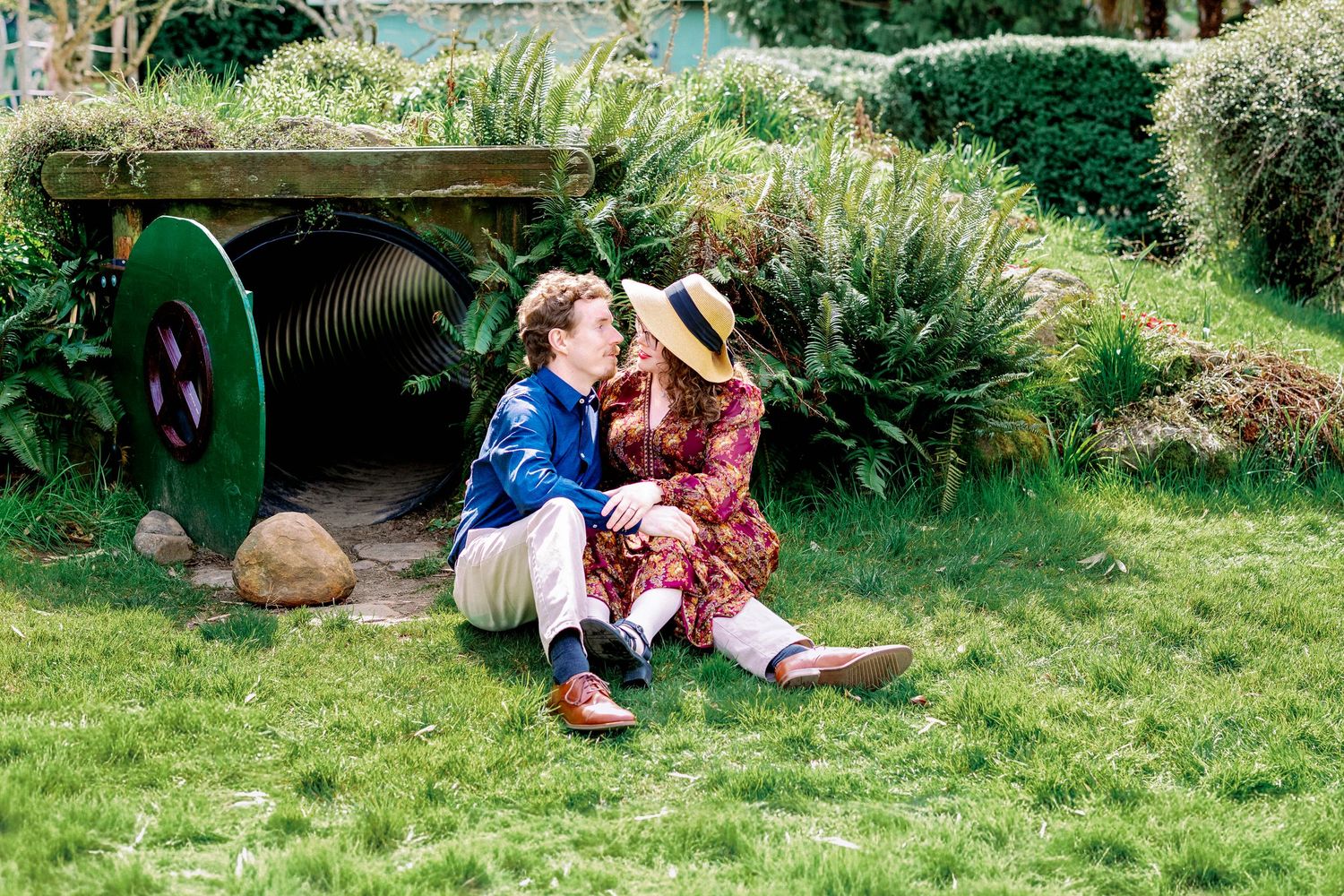 Two people sit together on green grass in a garden setting with a wooden structure and plants in the background.
