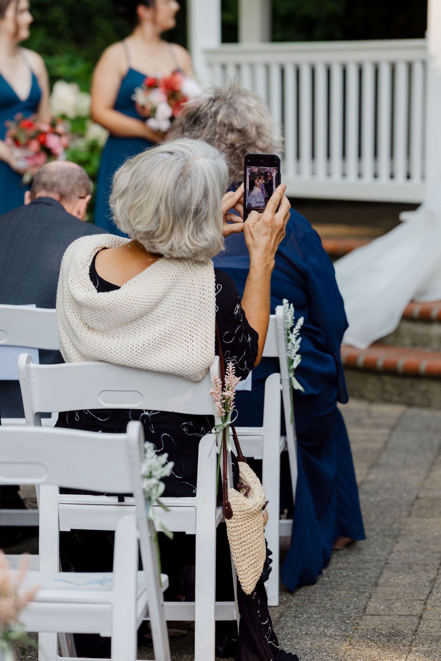 Two elderly women seated at outdoor wedding ceremony photograph the bride on smartphones.