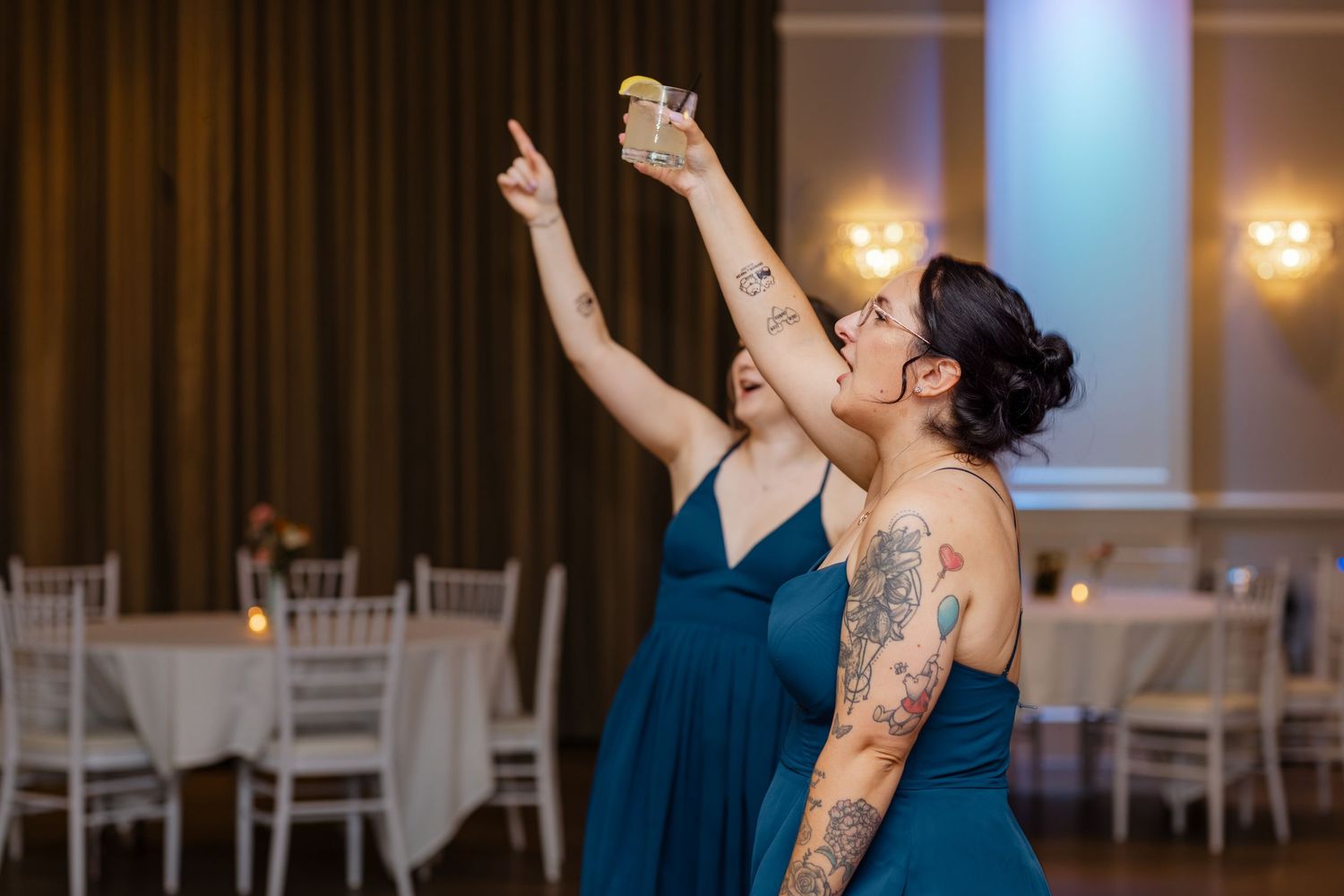 Two bridesmaids in teal dresses with tattoos celebrate joyfully on dance floor at wedding reception.