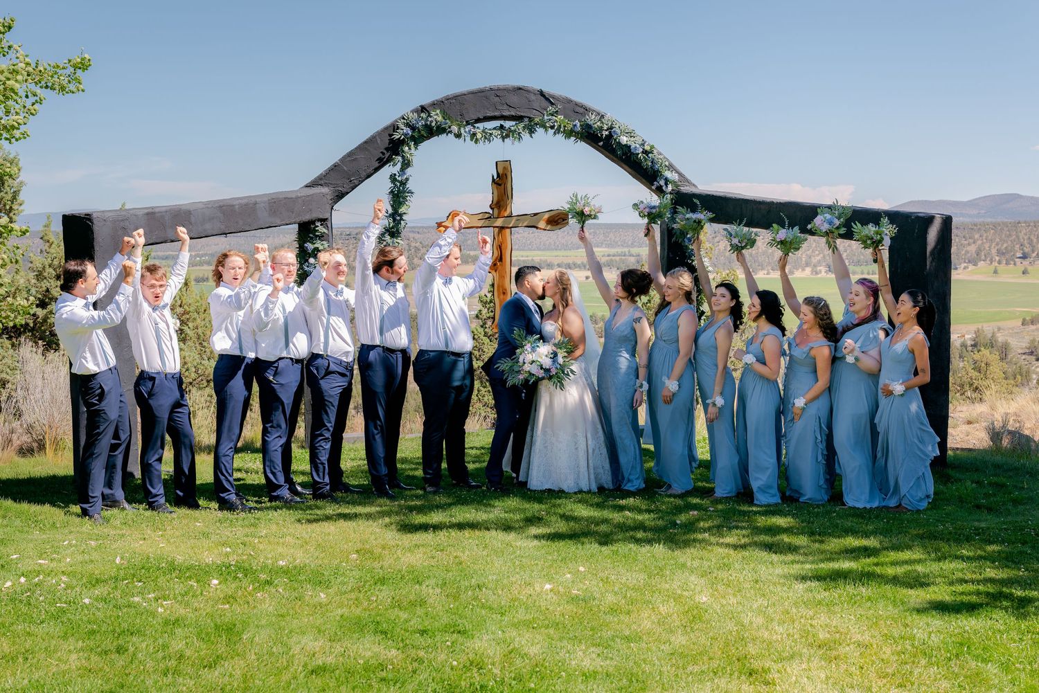Wedding party celebrates in front of floral arch with bridesmaids in blue and groomsmen in navy.