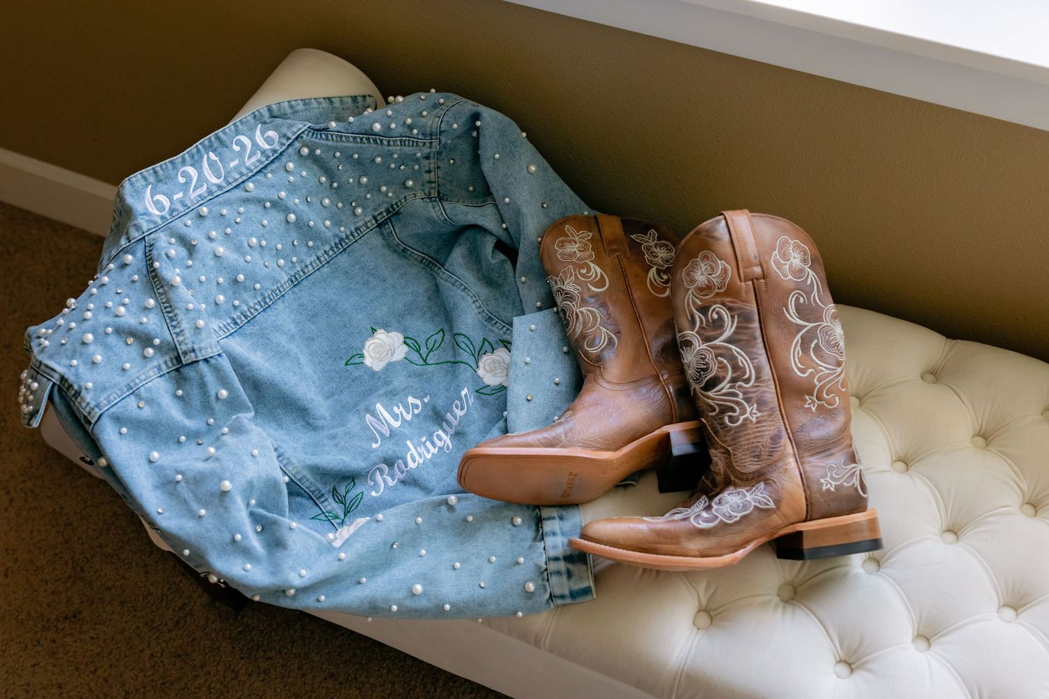 Bedazzled denim bridal jacket reading Mrs. with brown cowboy boots on tufted bench.
