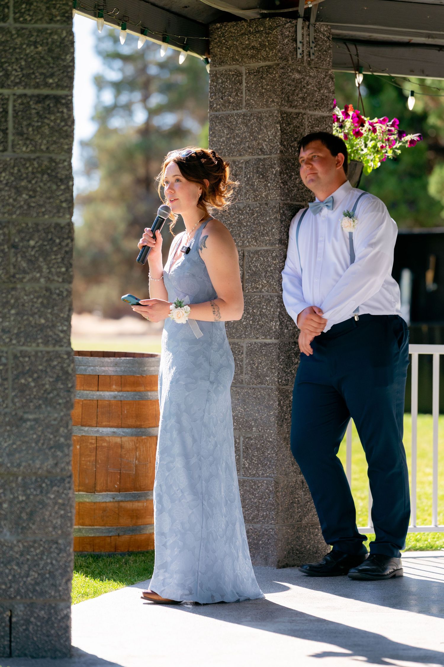 Bridesmaid in a grey lace gown speaks into a microphone during a wedding toast beside the groom outdoors.