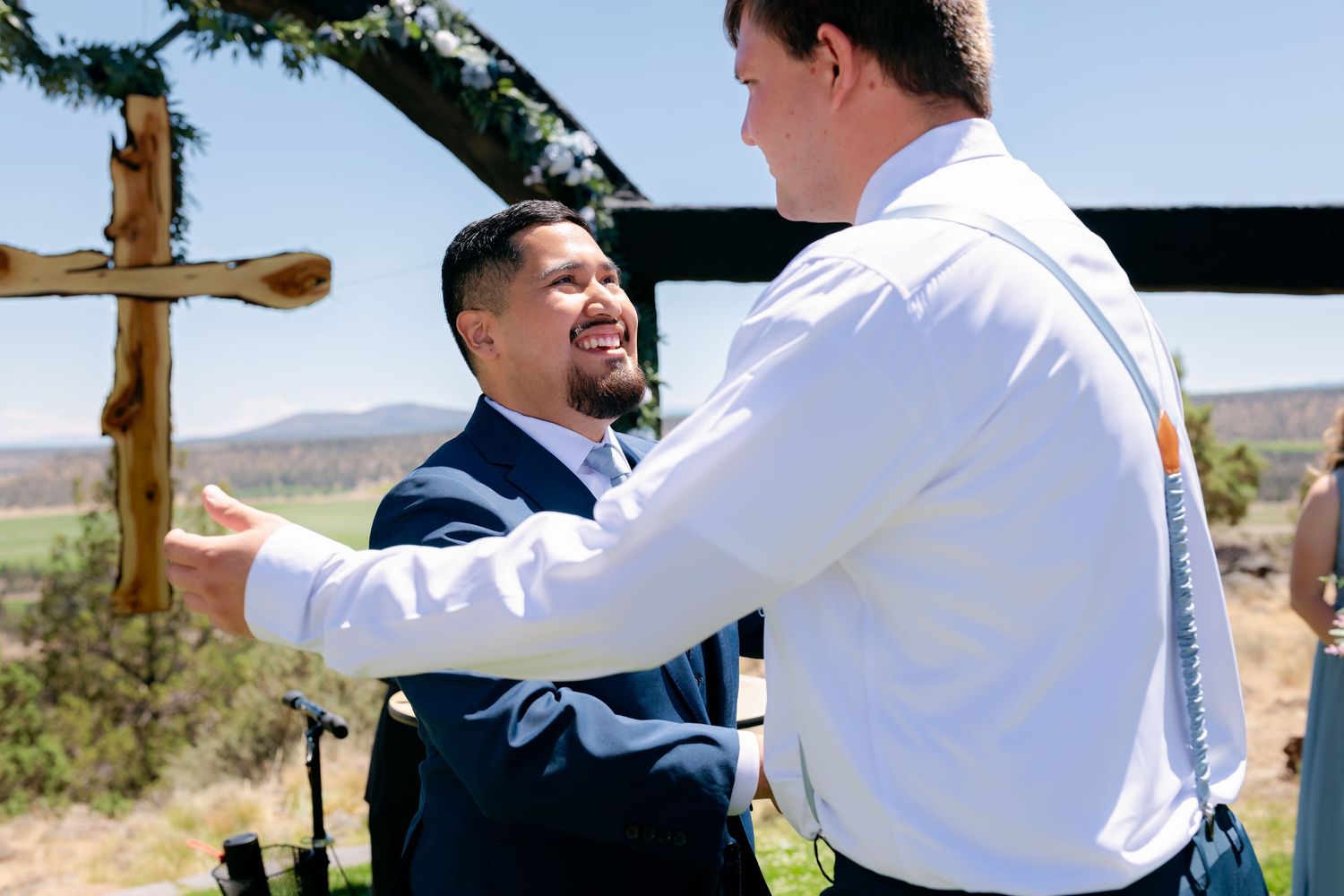 Groom in navy suit shares a joyful embrace with groomsman at outdoor wedding ceremony.