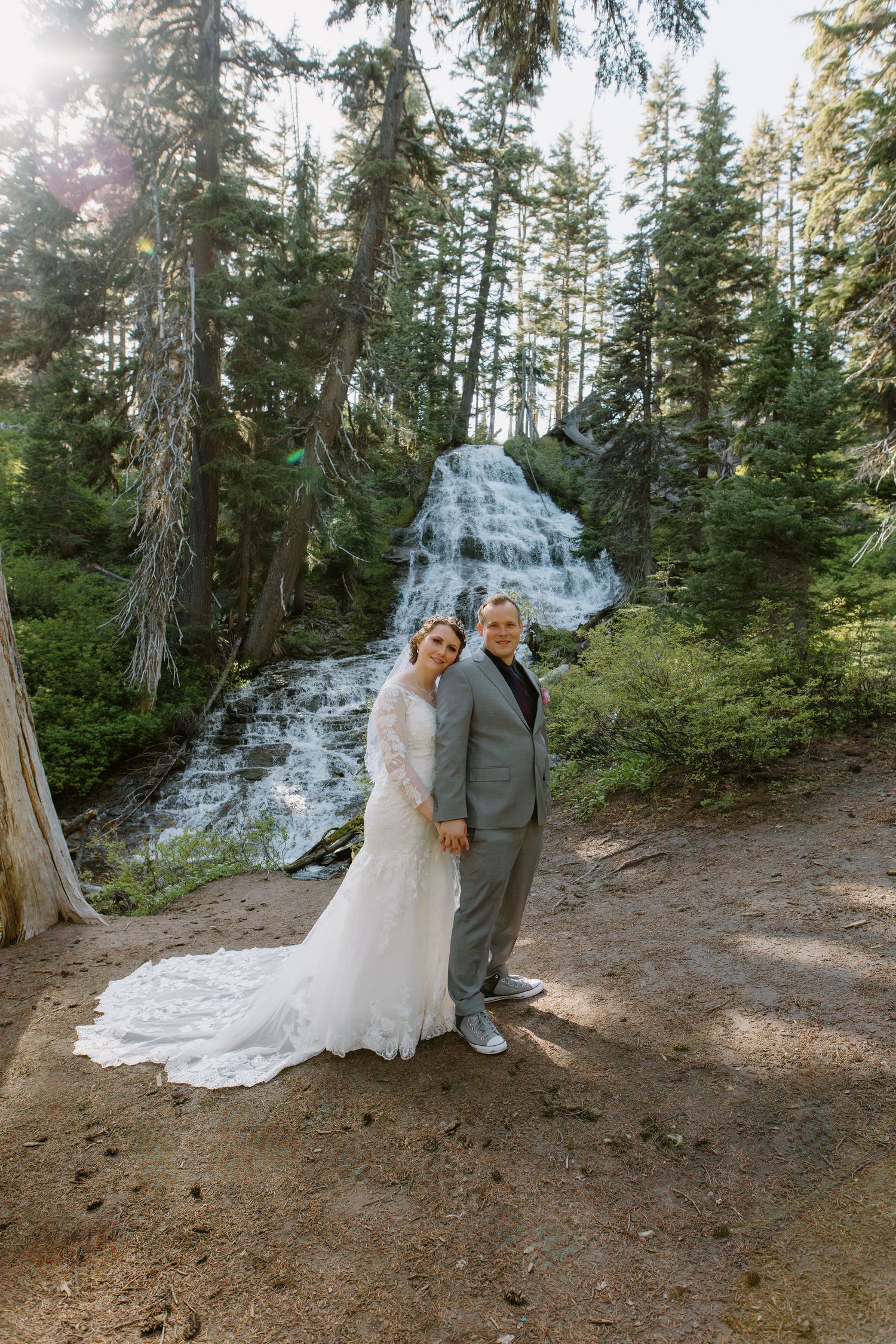 Bride and groom pose together in front of scenic waterfall surrounded by tall evergreen trees.