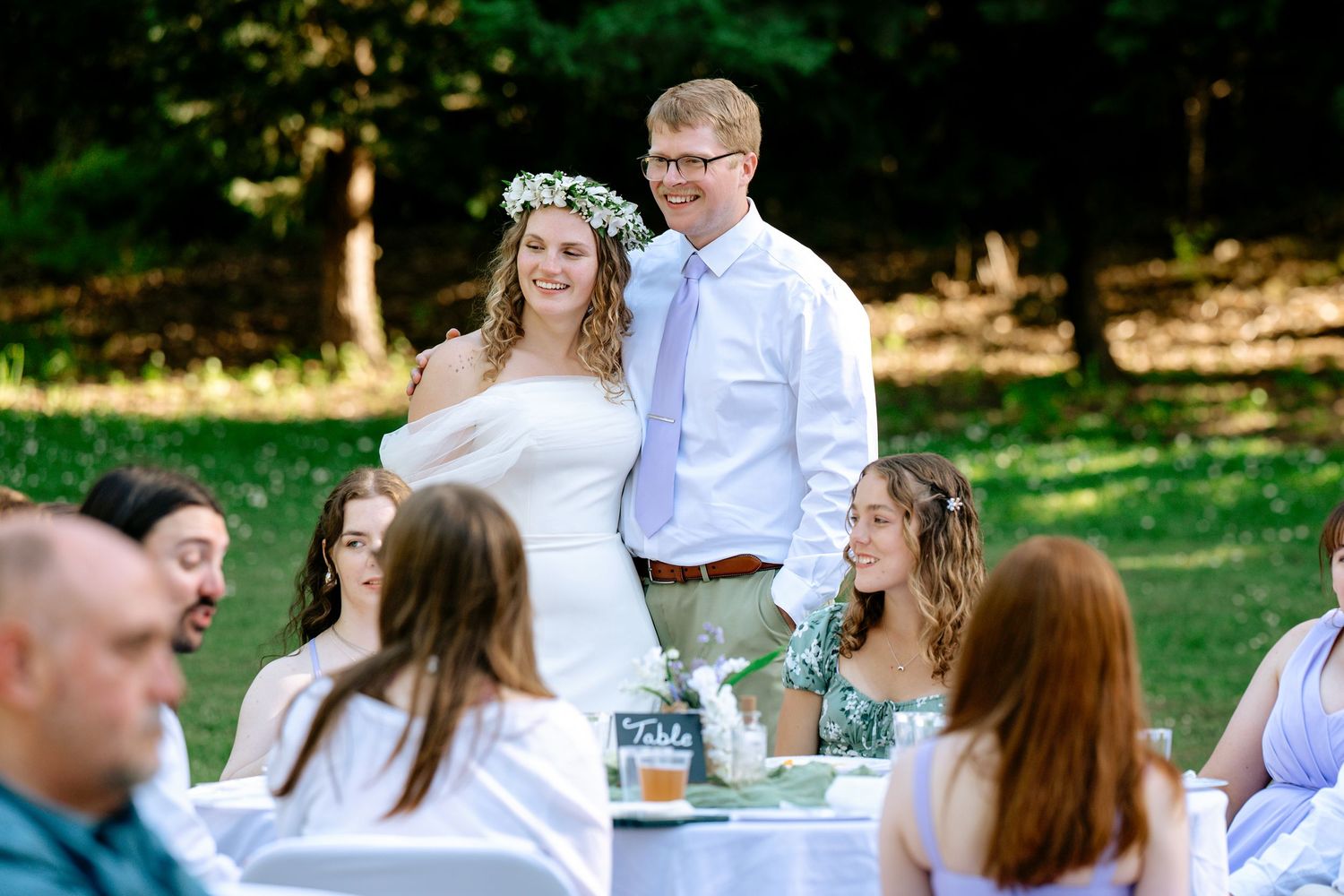 Bride wearing floral crown and groom in blue tie smile at guests seated at outdoor wedding reception tables.