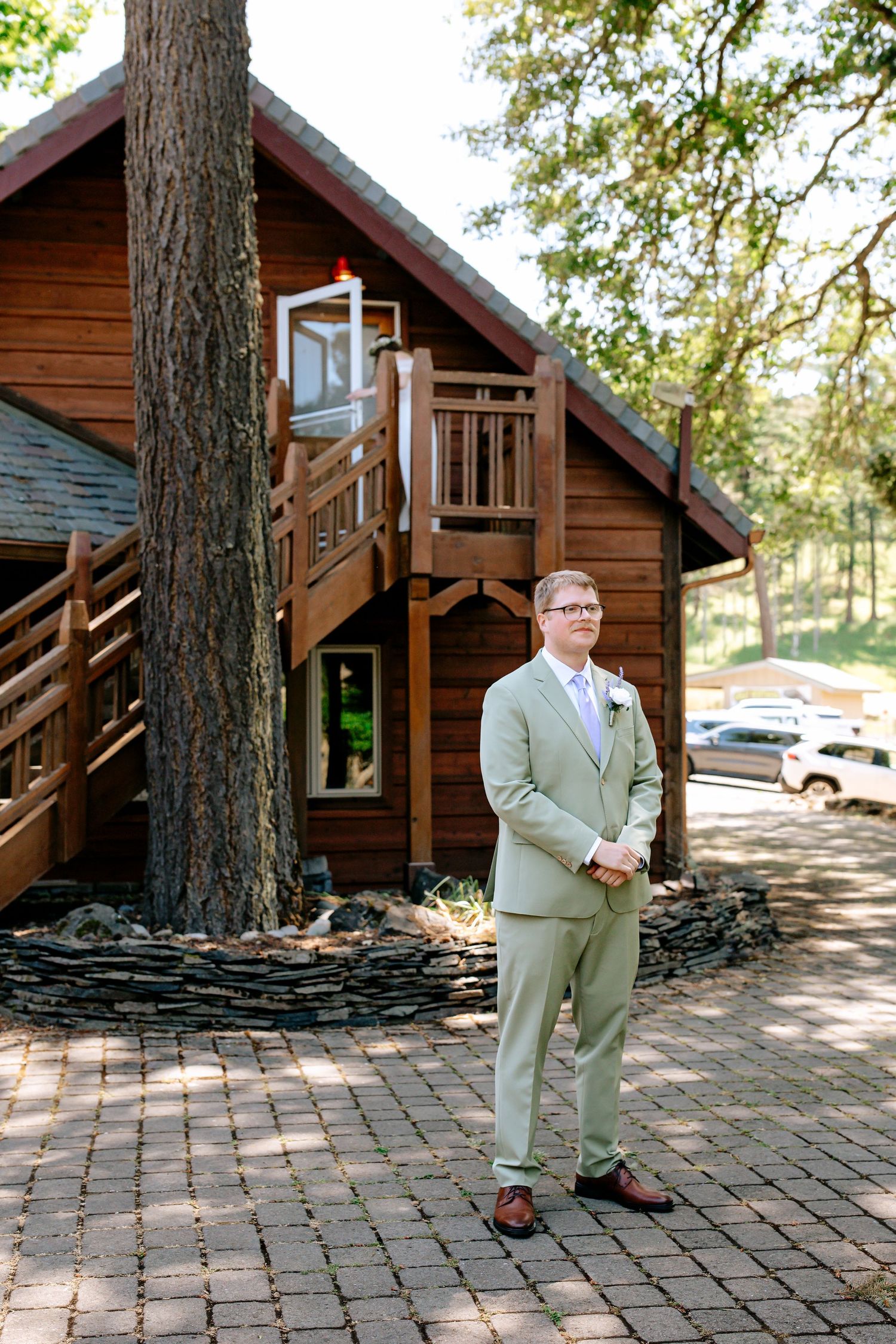 Groom in sage green suit standing on brick path outside rustic wooden lodge venue on wedding day.