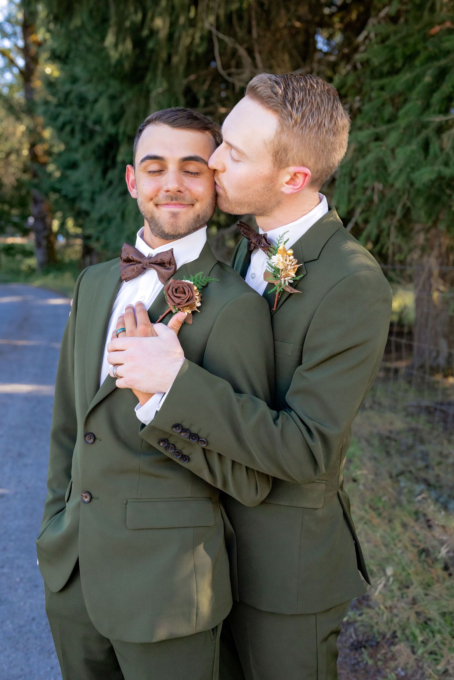Two grooms in olive green suits with bow ties share a tender kiss on their wedding day outdoors.