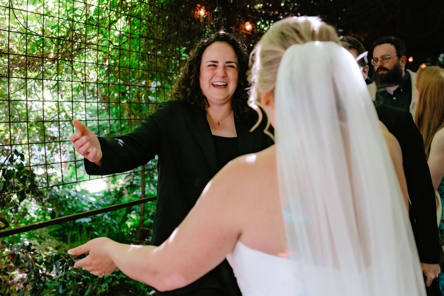 Wedding officiant in black jacket gestures warmly to a bride in white gown and veil at an elegant venue.