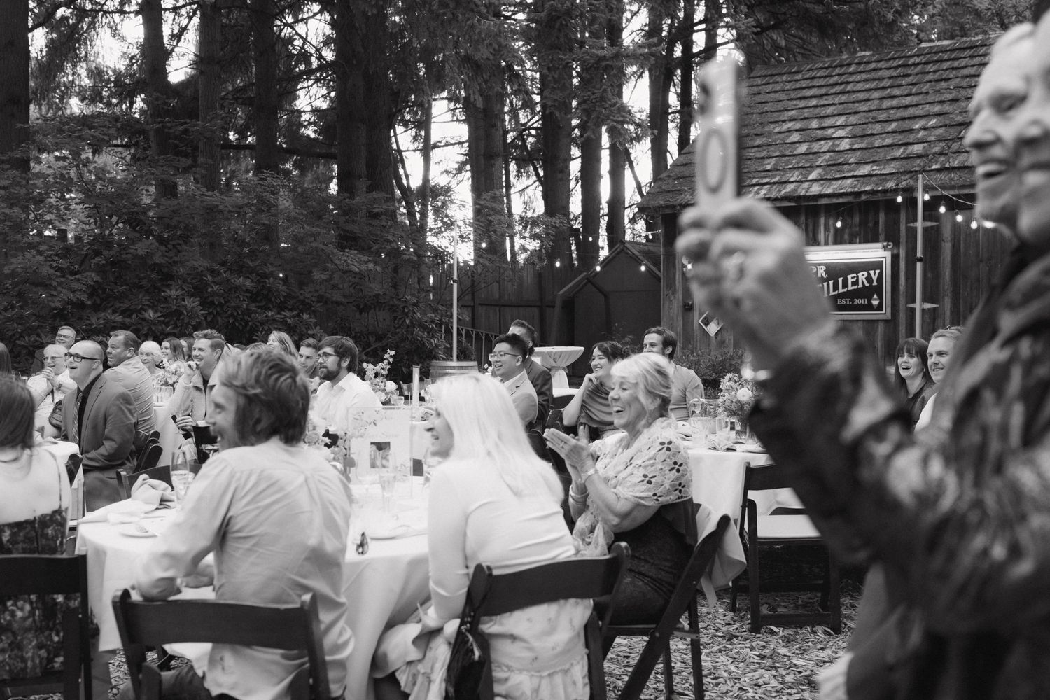 Guests celebrate at outdoor wedding reception dinner under tall trees.