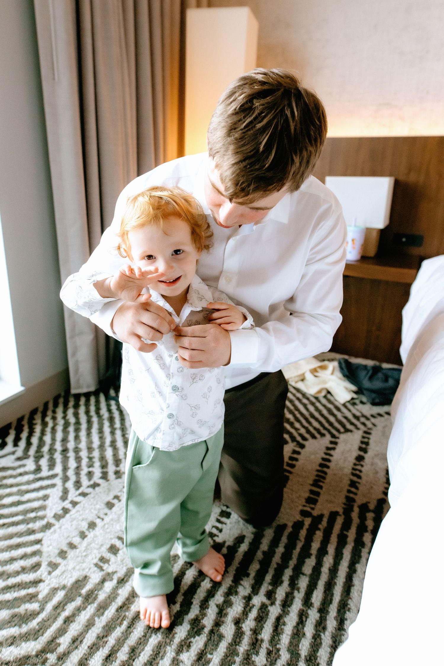 Older brother helps young redhead toddler button white dress shirt in a hotel room before a wedding.