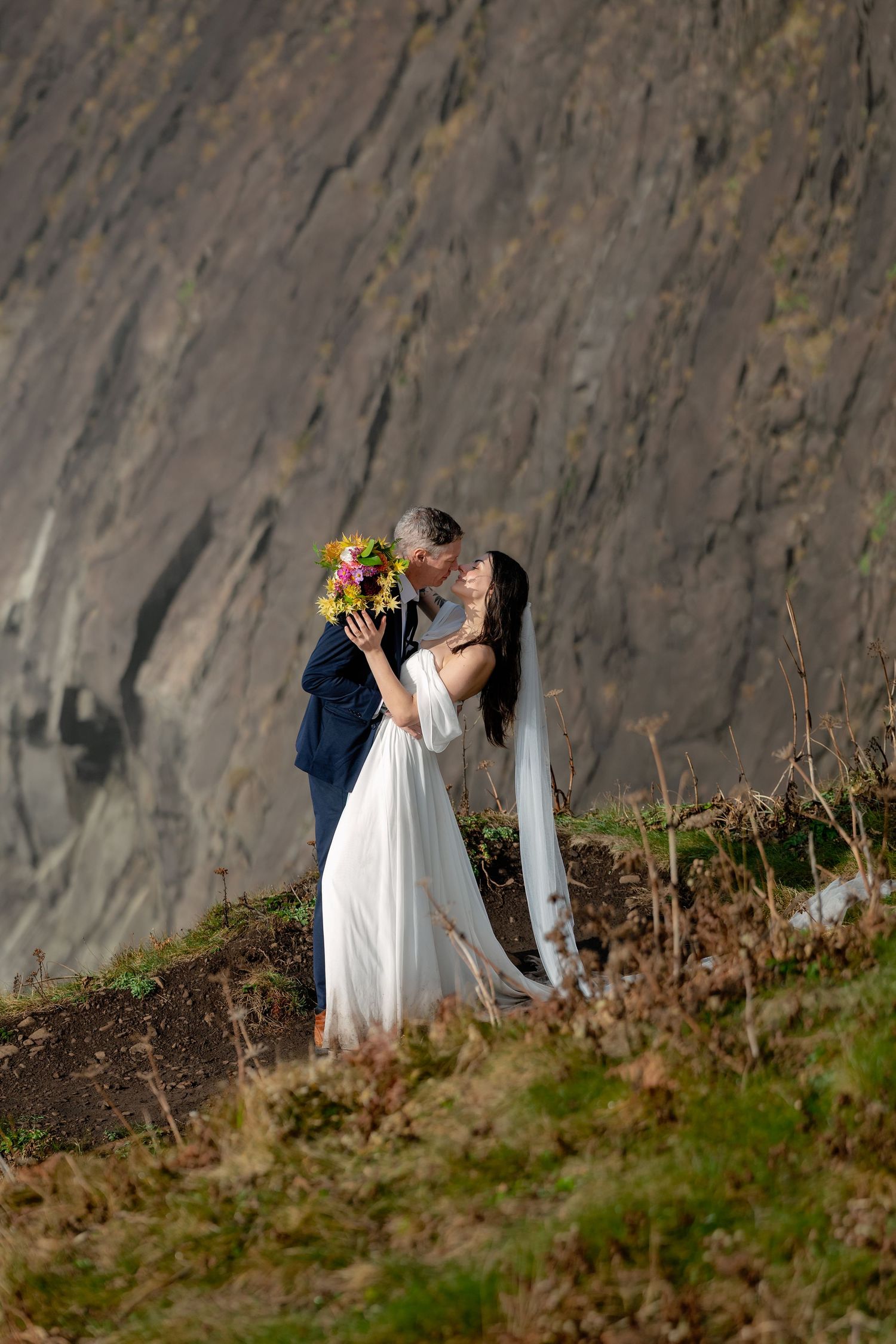 A couple in wedding attire share a romantic moment against a dramatic cliff backdrop.