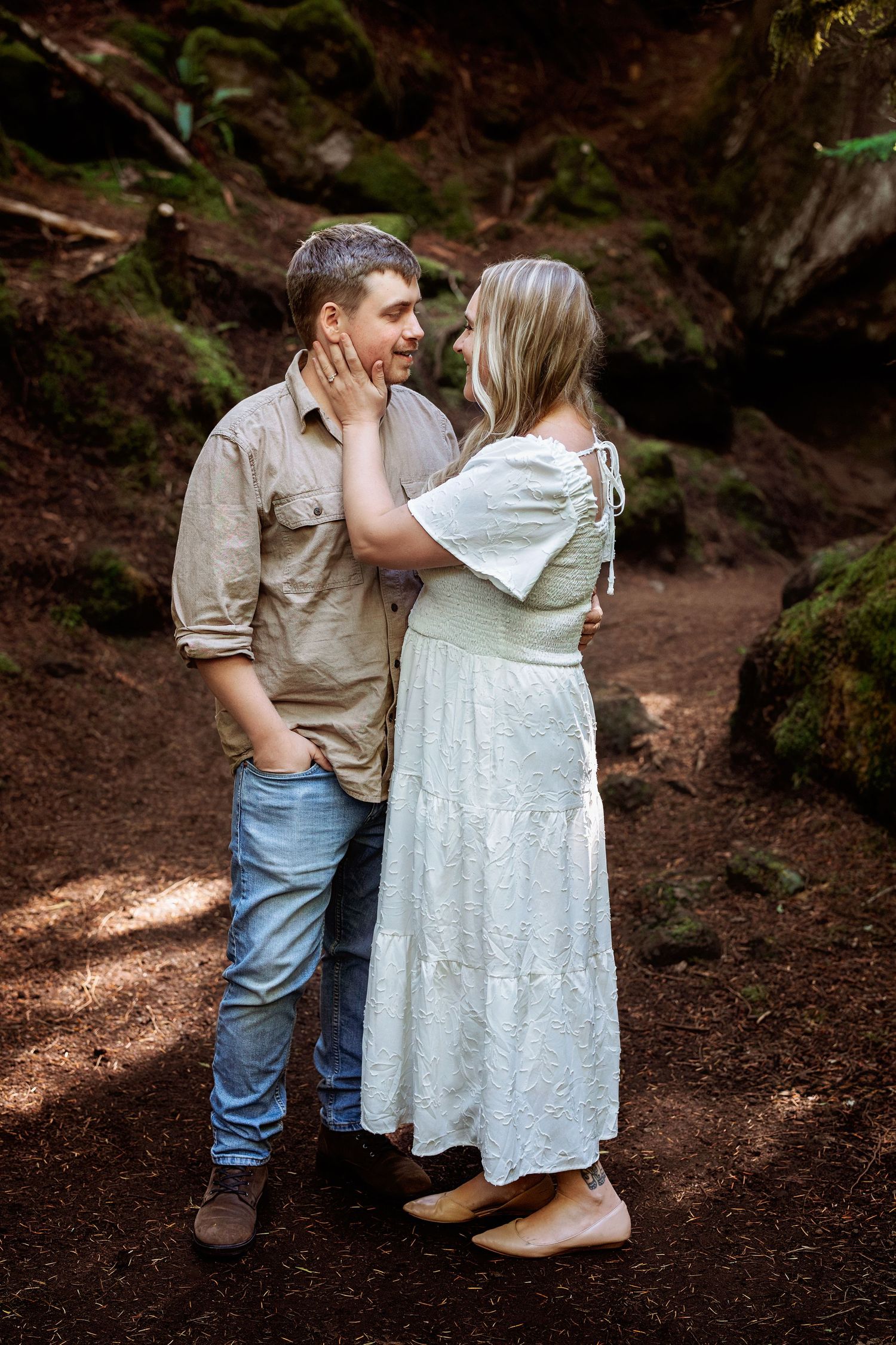 A couple shares an intimate moment together on a forest trail wearing casual attire.