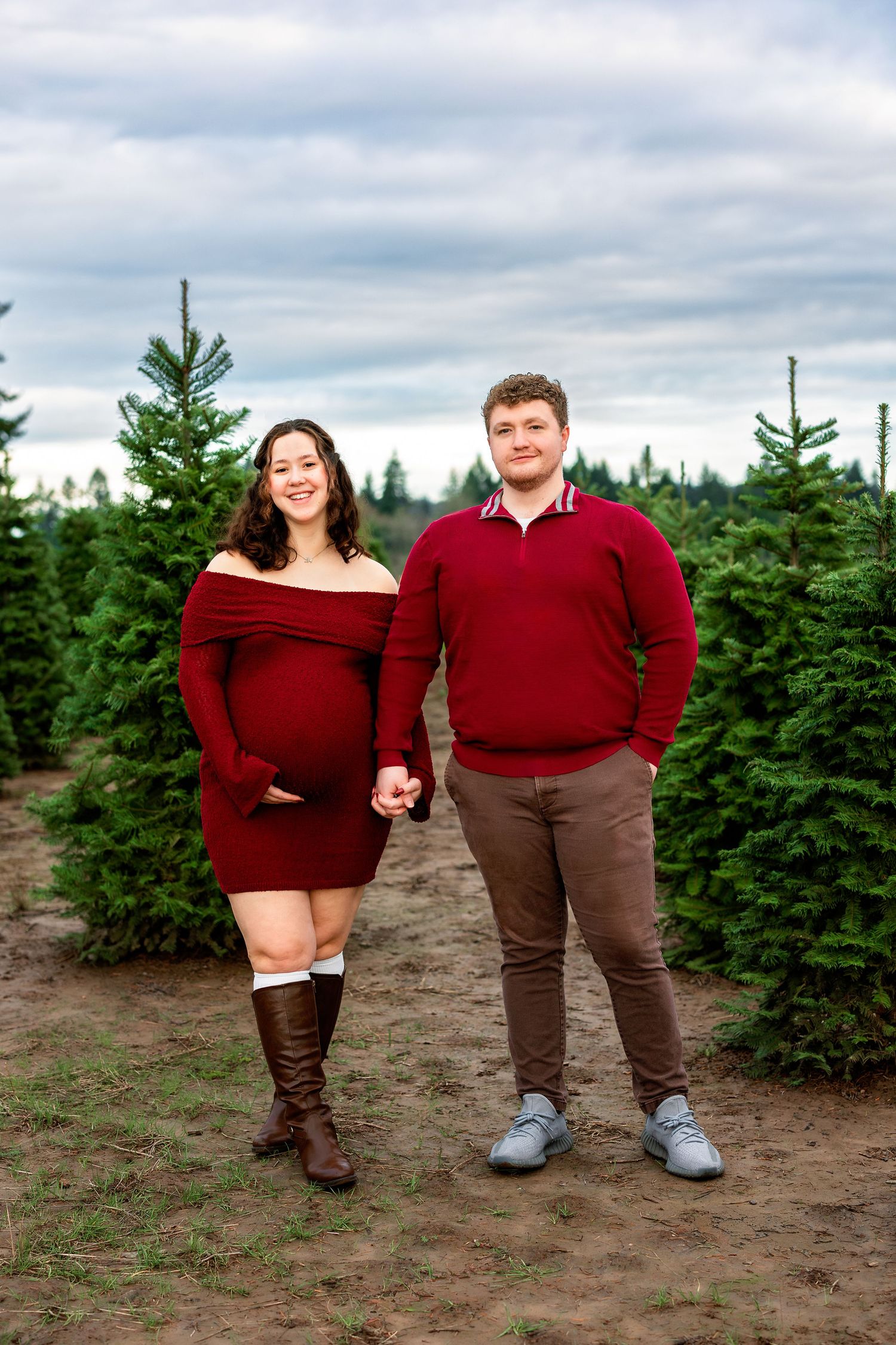 Couple in matching burgundy sweaters pose together at a Christmas tree farm on a cloudy winter day.