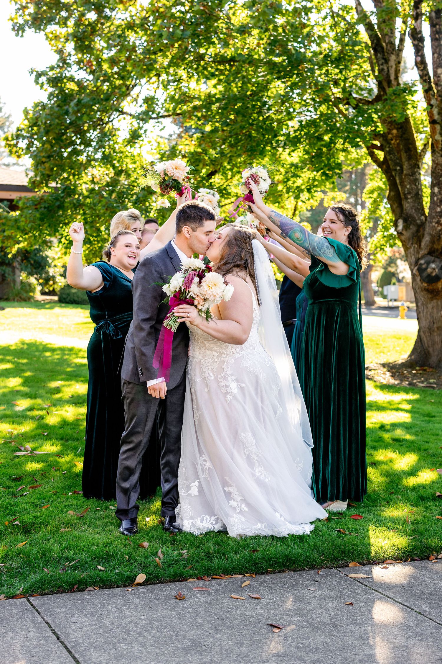 Wedding party celebrates on a sunny lawn with joyful raised arms and cheering.