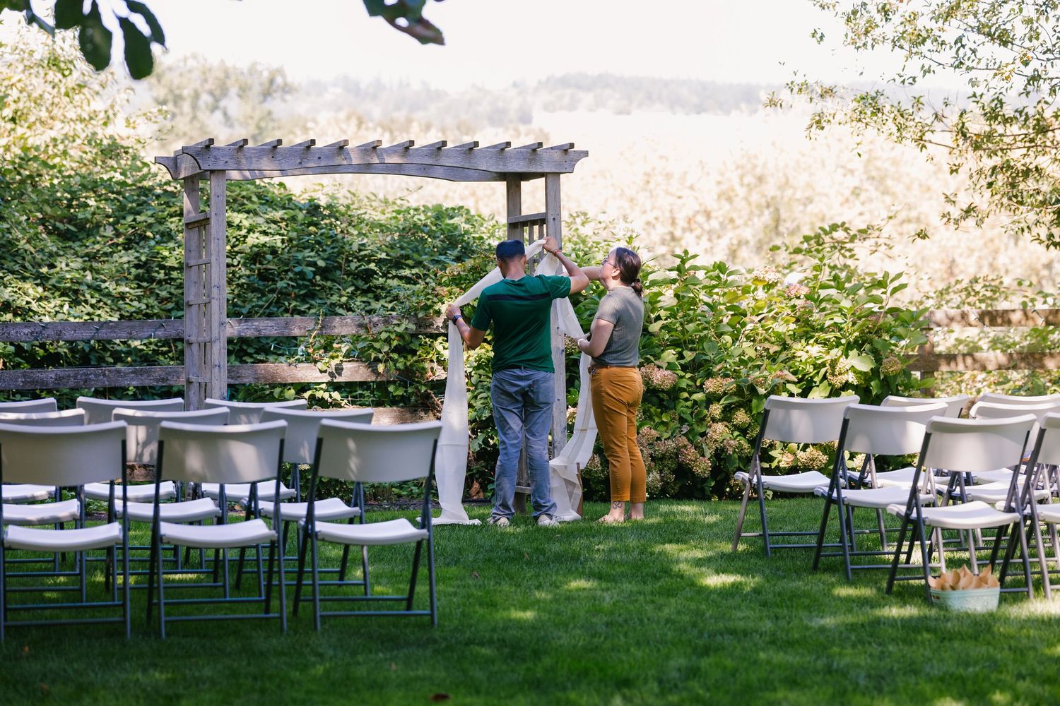A series of outdoor wedding reception tables and chairs arranged under trees with a pergola visible in the background.