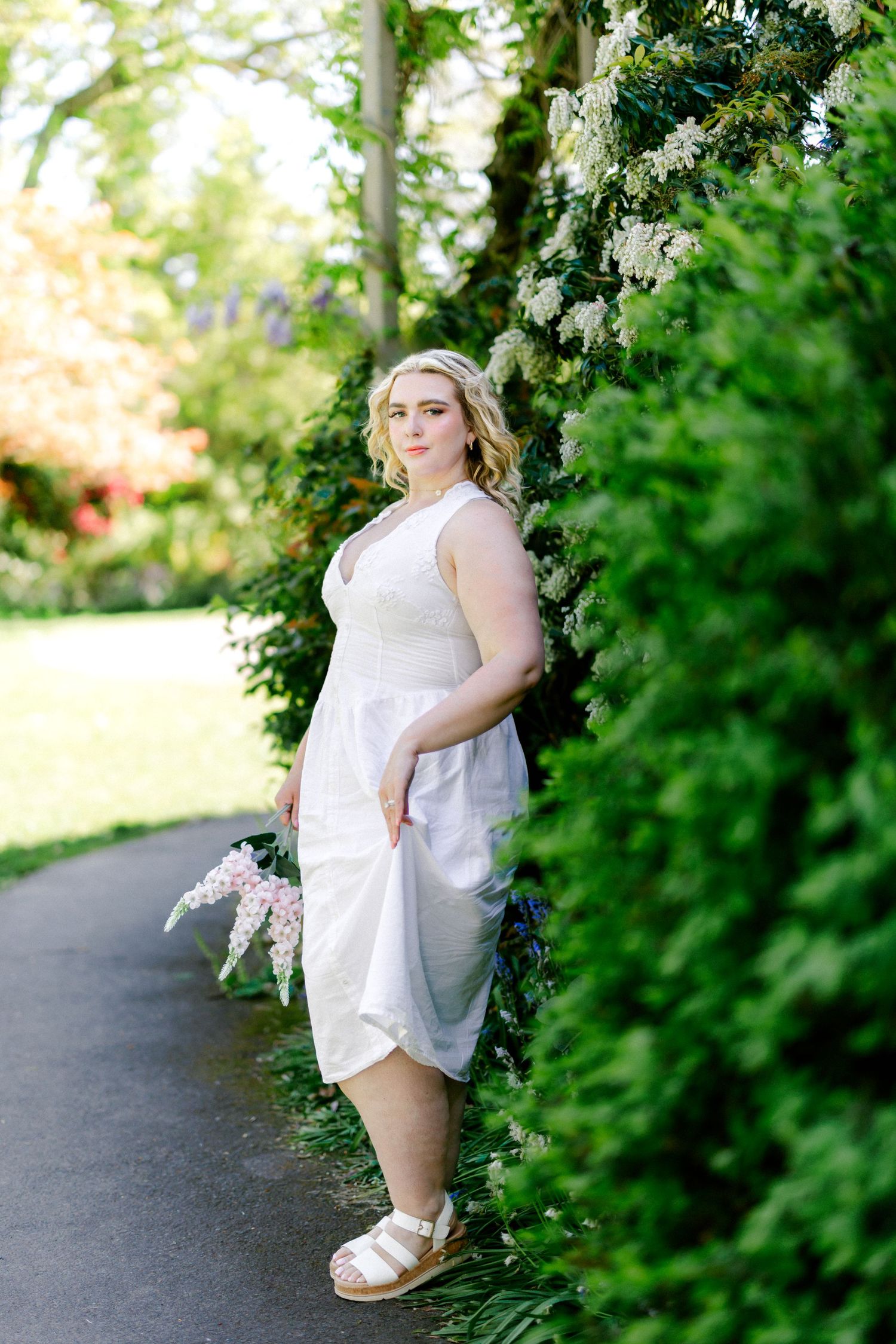 Blonde woman in white midi dress and sandals posing by lush green hedge in sunny garden.