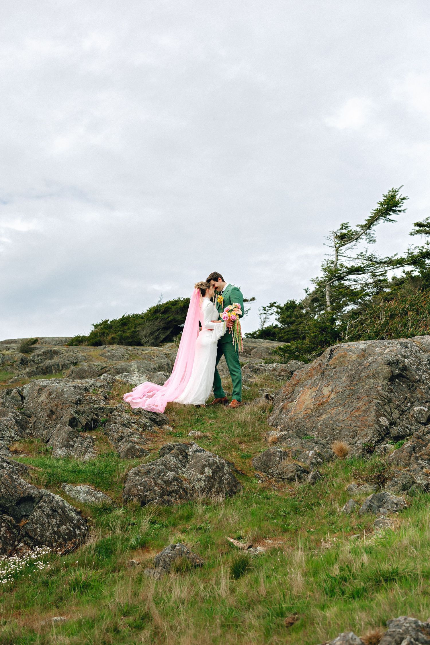 Wedding couple embracing on rocky coastal terrain, groom in green suit holding bride in white dress with flowing pink cape.