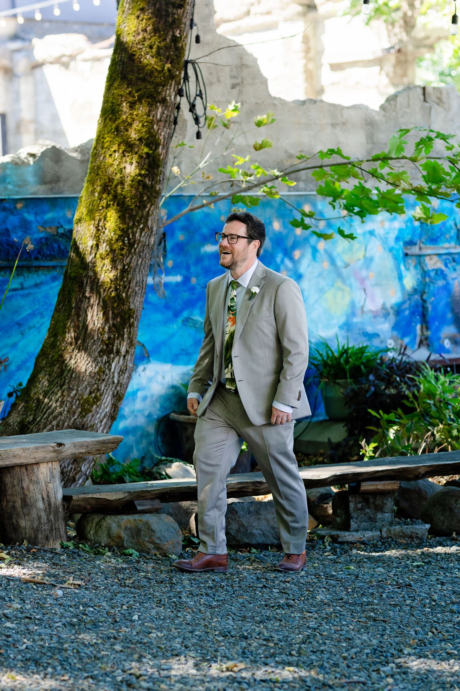 Groom in tan suit with floral tie stands confidently in garden courtyard with blue mural backdrop.