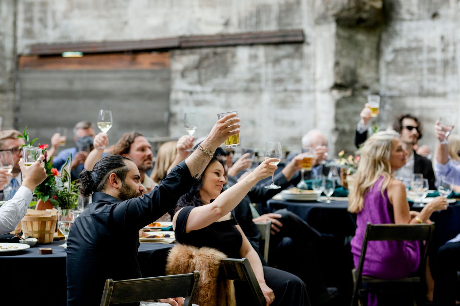Wedding guests raise glasses in a toast during outdoor reception at rustic stone venue.