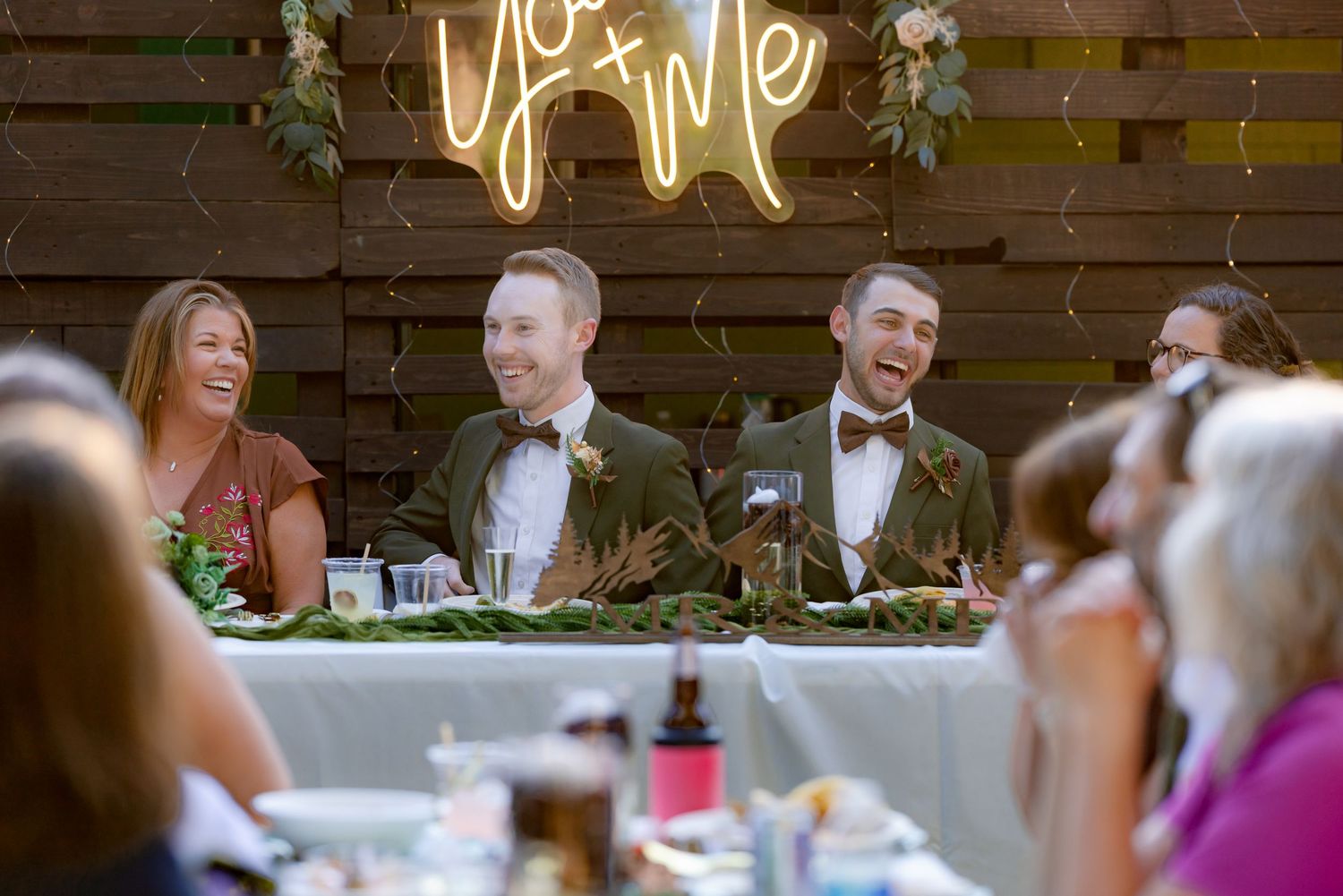 Two grooms in green velvet suits laugh joyfully at their sweetheart table under a neon You and Me sign.