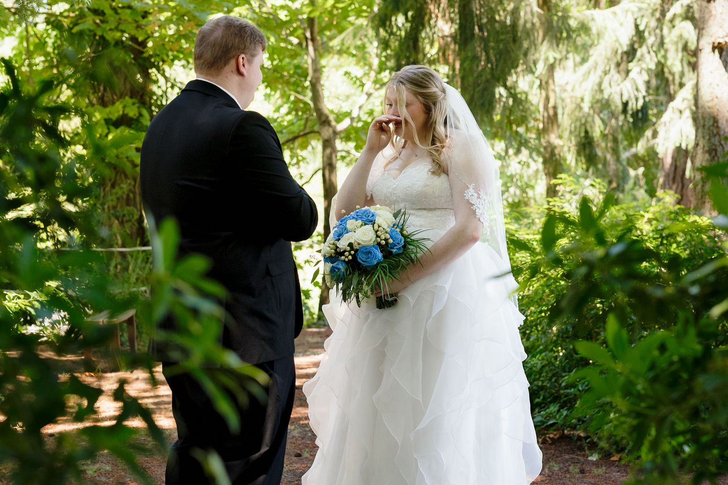 Bride and groom share emotional first look moment in lush garden, bride holding blue and white bouquet wearing white gown.