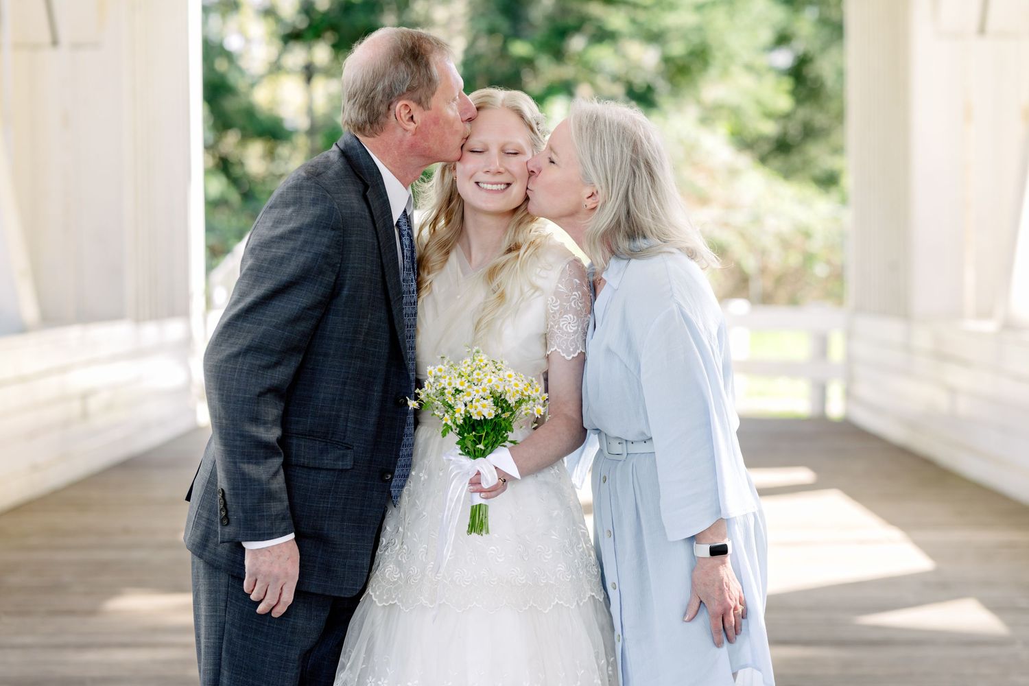 Bride in white lace gown holding yellow bouquet receives kisses on cheeks from parents at outdoor wedding venue.