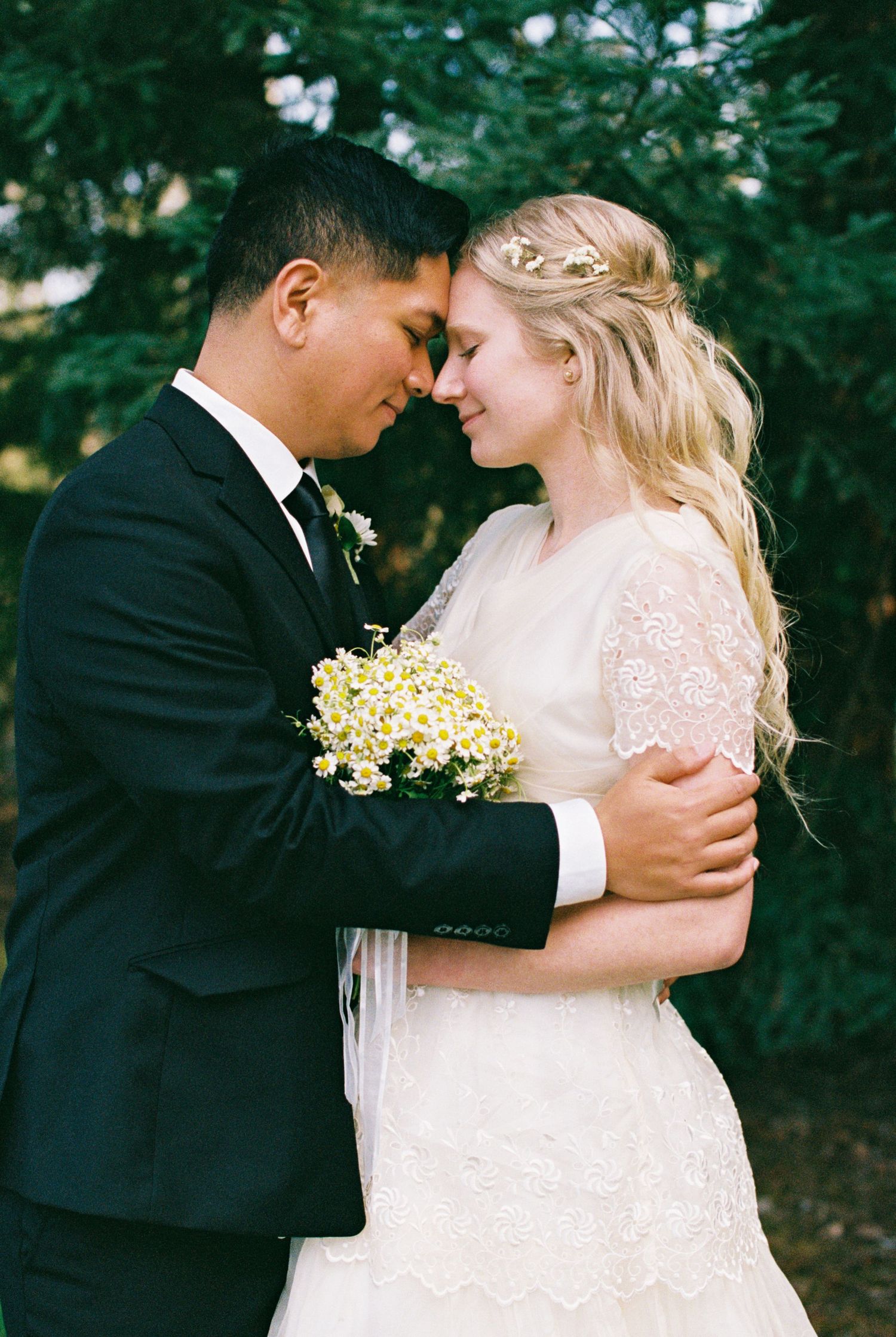 Bride and groom in wedding attire embrace forehead-to-forehead, holding yellow wildflowers outdoors.