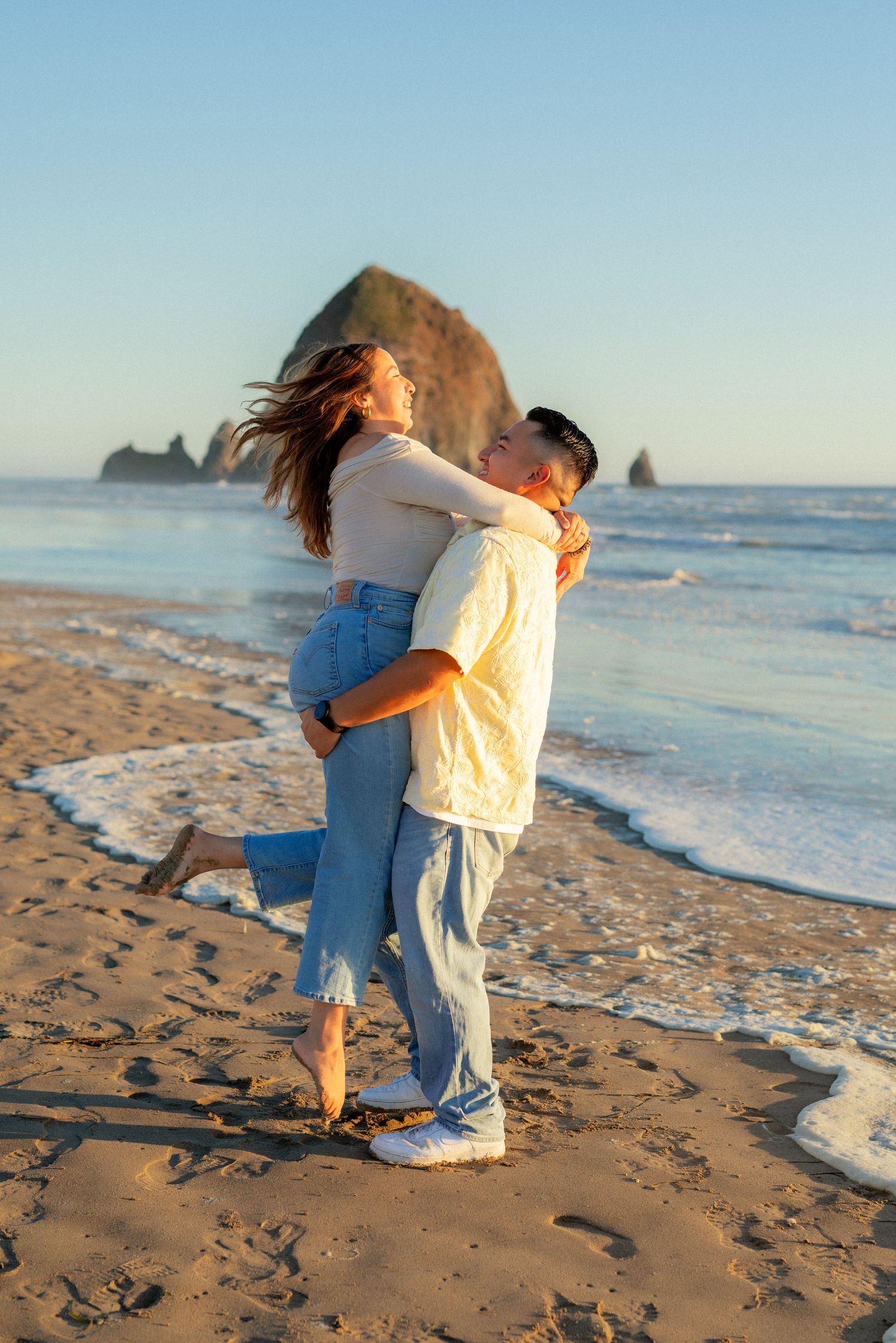 Couple embracing on Cannon Beach at golden hour with Haystack Rock in background.