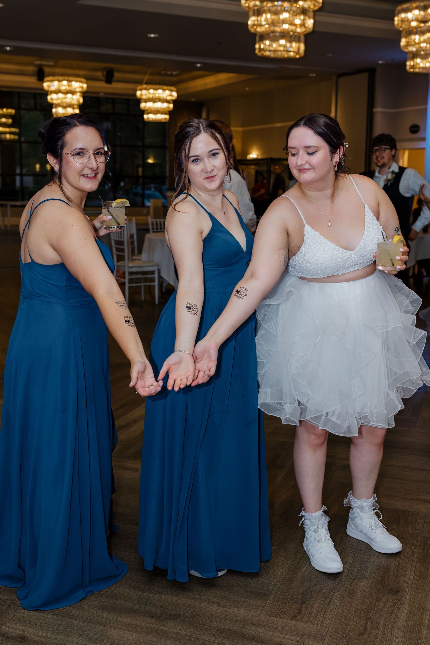 Three women in blue bridesmaid dresses and white bridal outfit showing matching tattoos on their arms.