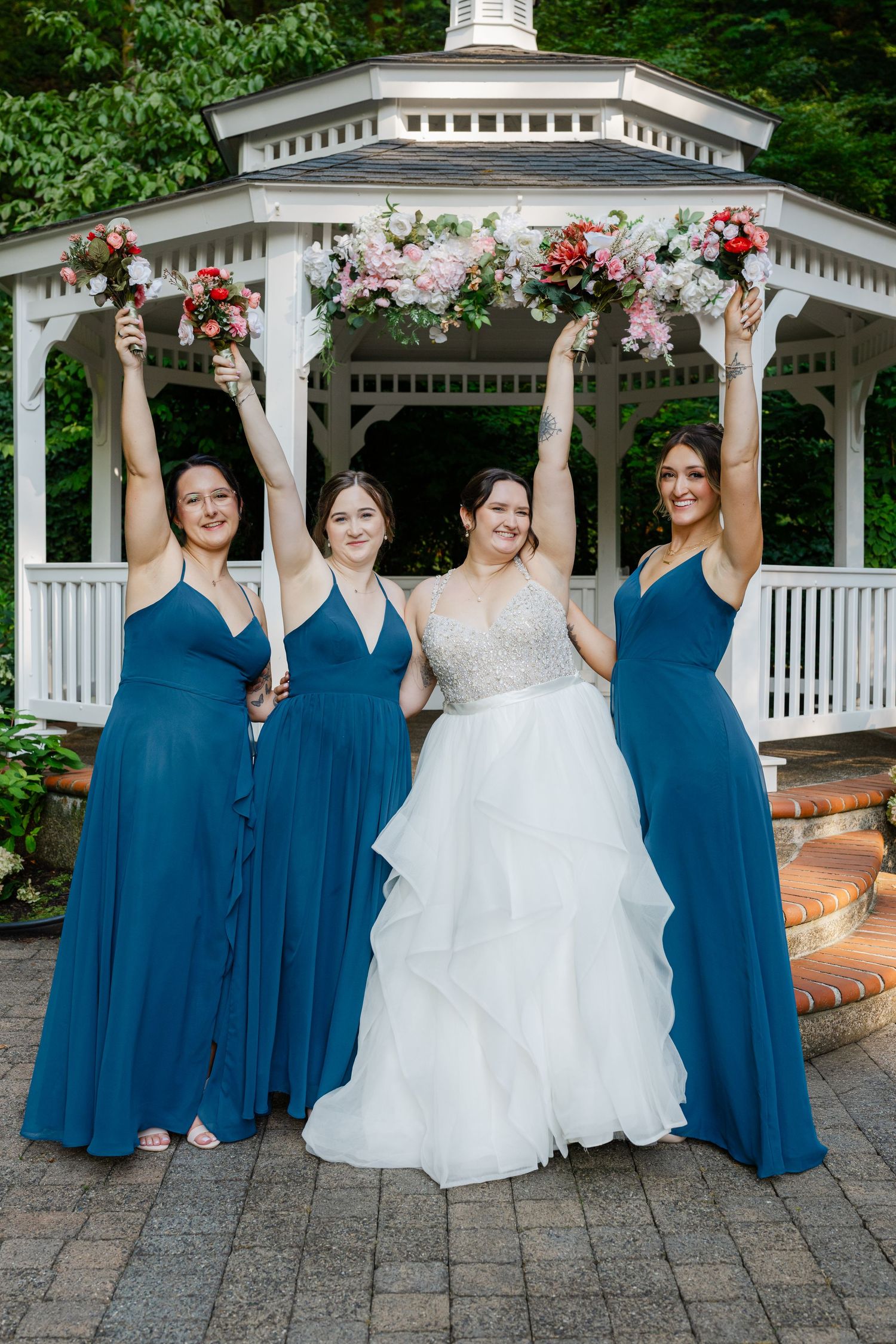 Bride and three bridesmaids in blue dresses raise bouquets in front of a white gazebo.