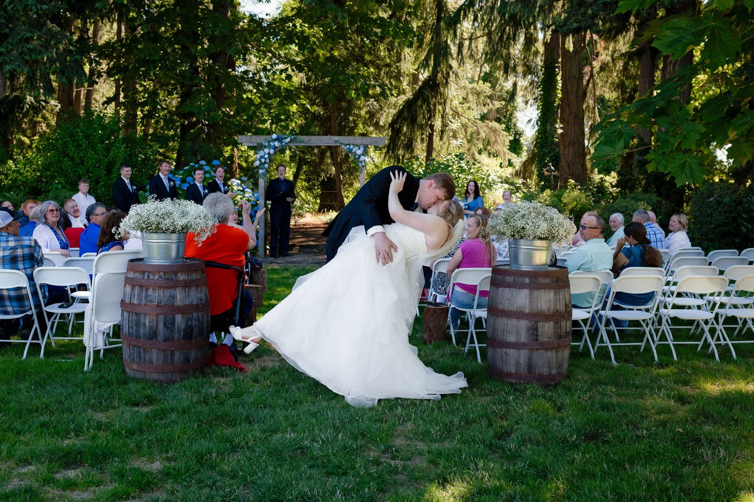 Groom dips bride in white dress for romantic kiss at outdoor garden wedding reception.