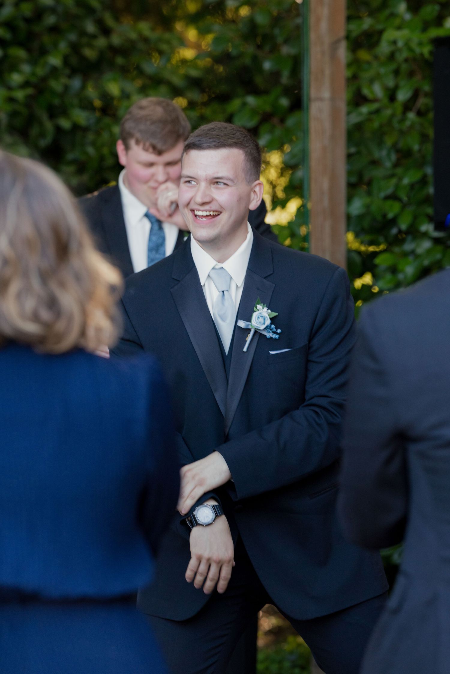 Smiling groomsman in black tuxedo with floral boutonniere laughs while greeting guests at outdoor wedding reception.